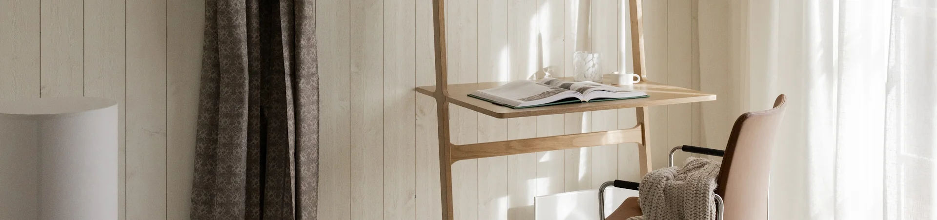 Een houten standaard met een open boek, glazen vaas en witte mok, naast een bruine leren stoel met een beige deken, badend in zonlicht.