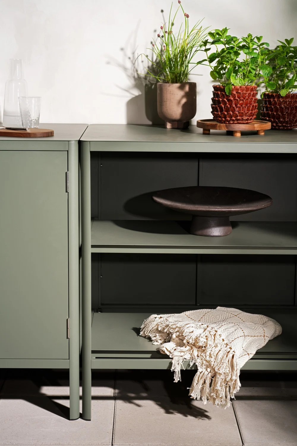 Salie groen dressoir en open kast met potplanten, een glazen fles, een decoratieve schaal en een textuurdeken.