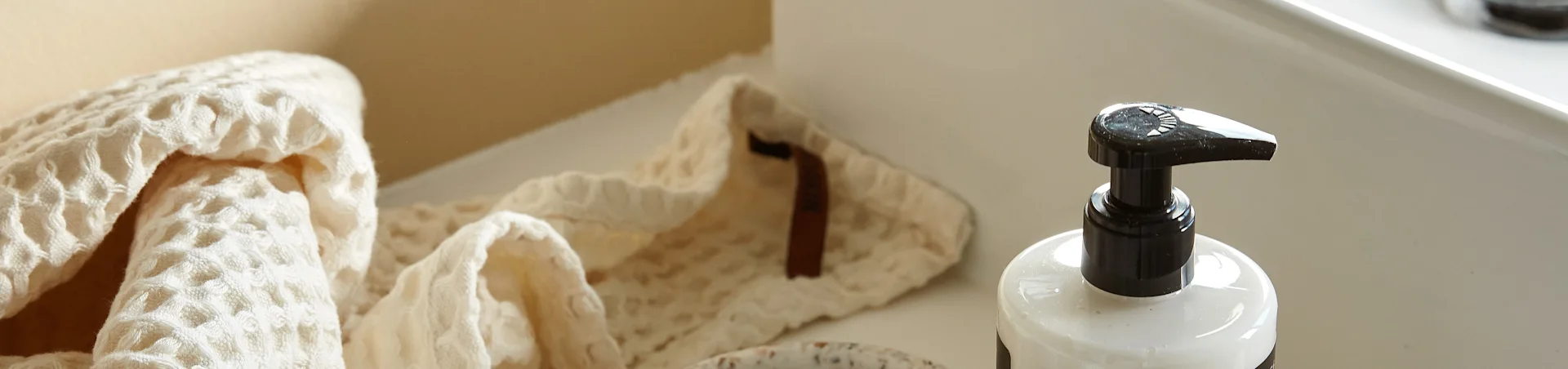 Een lichtgekleurde, wafelgeweven handdoek gevouwen naast een witte pompfles op een schoon, licht oppervlak.