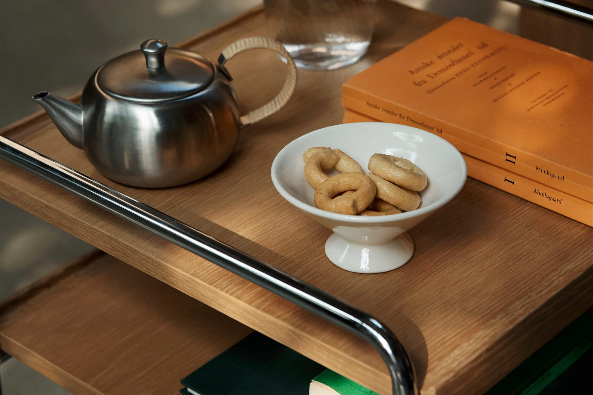 Een metalen theepot, witte kom met ringkoekjes en twee oranje boeken op een houten trolley.