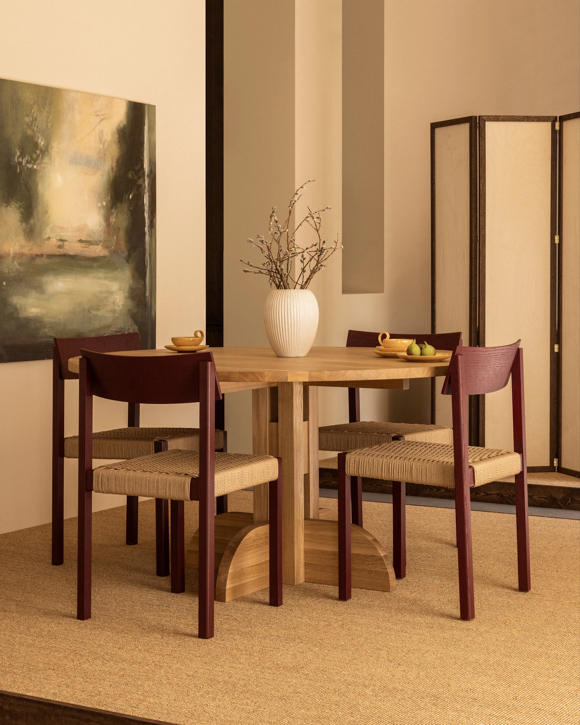 Een eetkamer met een ronde houten tafel, vier stoelen met roodbruine frames en gevlochten zittingen, en een vaas met wilgenkatjes.