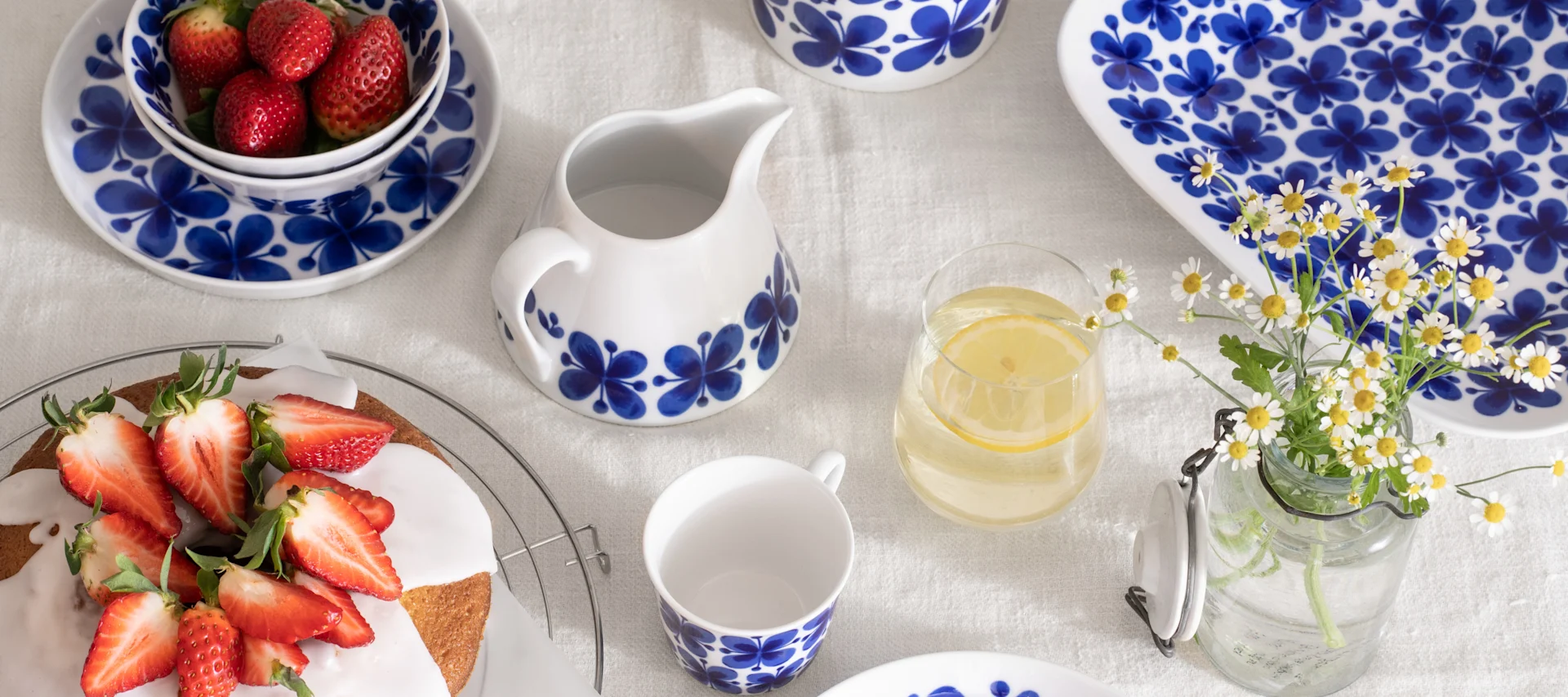 Tafel met wit- en blauwpatroon keramisch servies, een aardbeientaart, een kom aardbeien, citroenwater en madeliefjes.