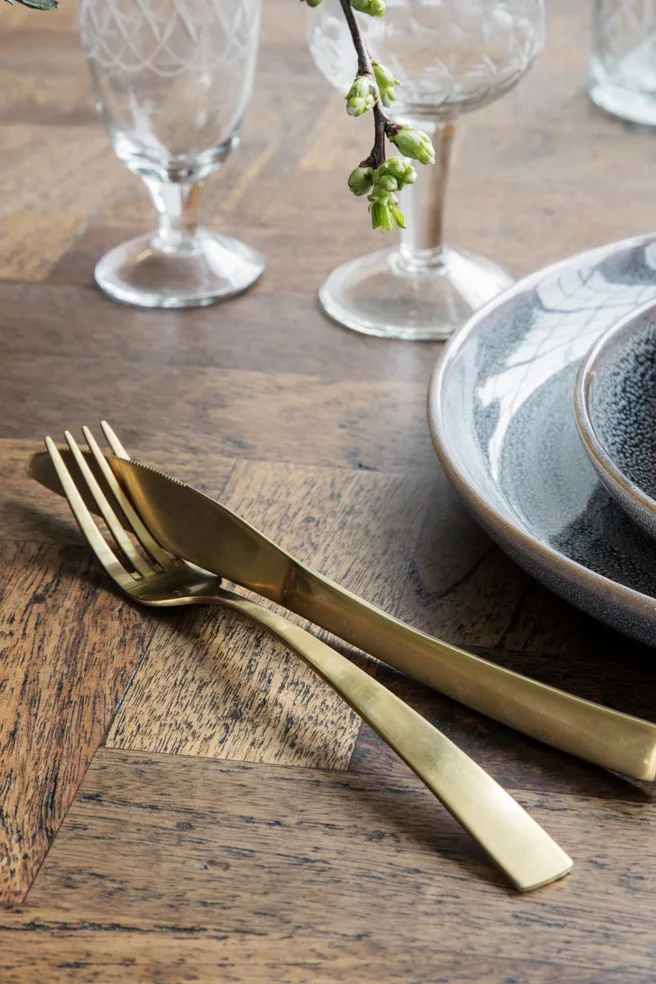 Gouden vork en mes op een houten tafel naast keramische borden en gedessineerde glazen, met een kleine tak met groene knoppen.