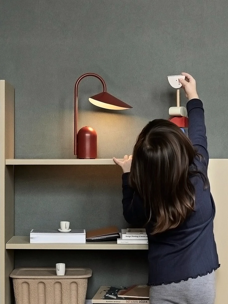 Een kind reikt naar een houten speelgoedvogel op een lichte plank met een rode lamp en boeken; een gevlochten mand staat op de vloer.