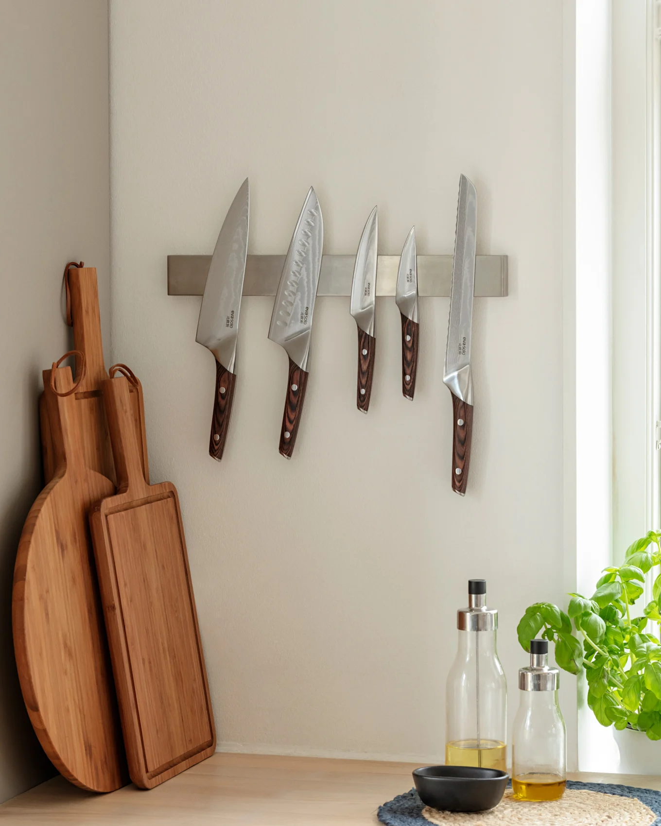 Keukenhoek met vijf messen aan een magneetstrip, twee houten snijplanken en twee flessen olijfolie op een houten aanrecht naast een basilicumplant.