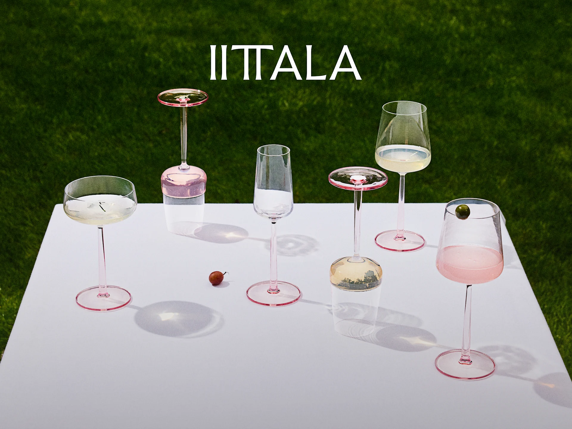 Iittala-logo boven een collectie glazen met roze steel op een witte tafel met groene grasachtergrond, sommige met drankjes.