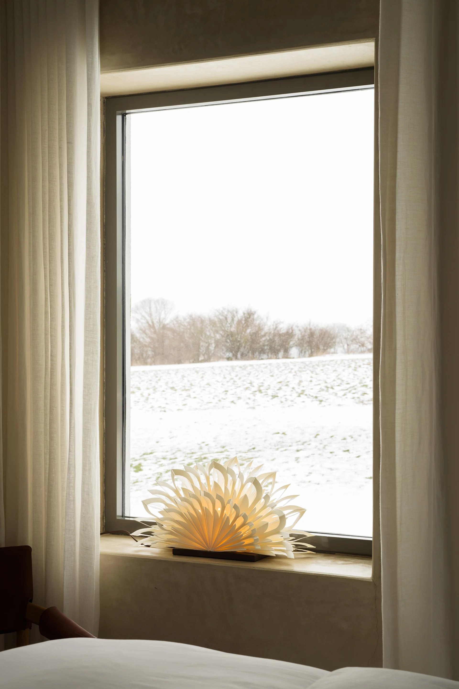 Raam met uitzicht op een besneeuwd veld, met witte gordijnen en een gloeiende papieren lamp op de vensterbank.