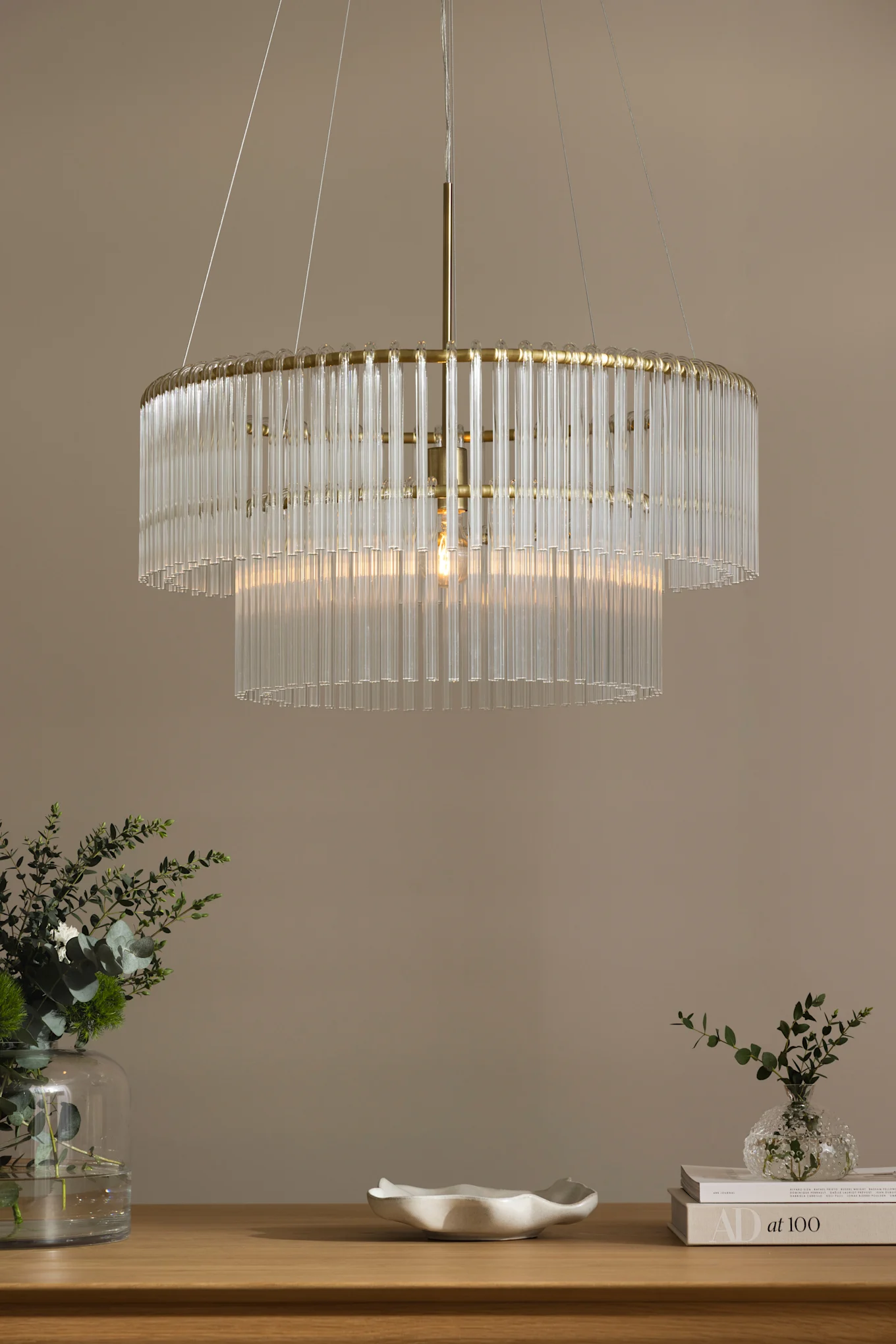 Twee-laags kroonluchter met heldere glazen staven en een goudkleurig frame, die een houten tafel met groene planten en een witte kom verlicht.