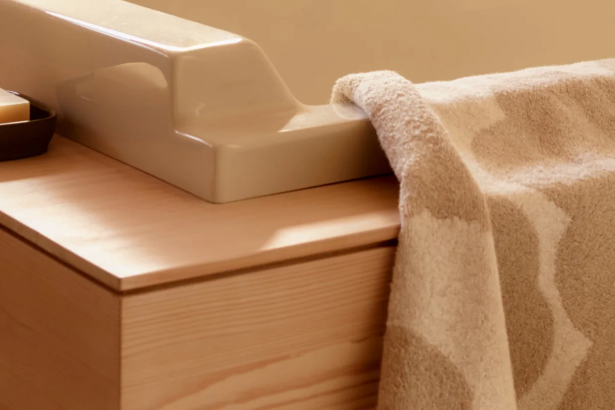 Close-up van een beige getextureerde handdoek gedrapeerd over een lichtgekleurd keramisch armatuur op een houten badmeubel met zeep.