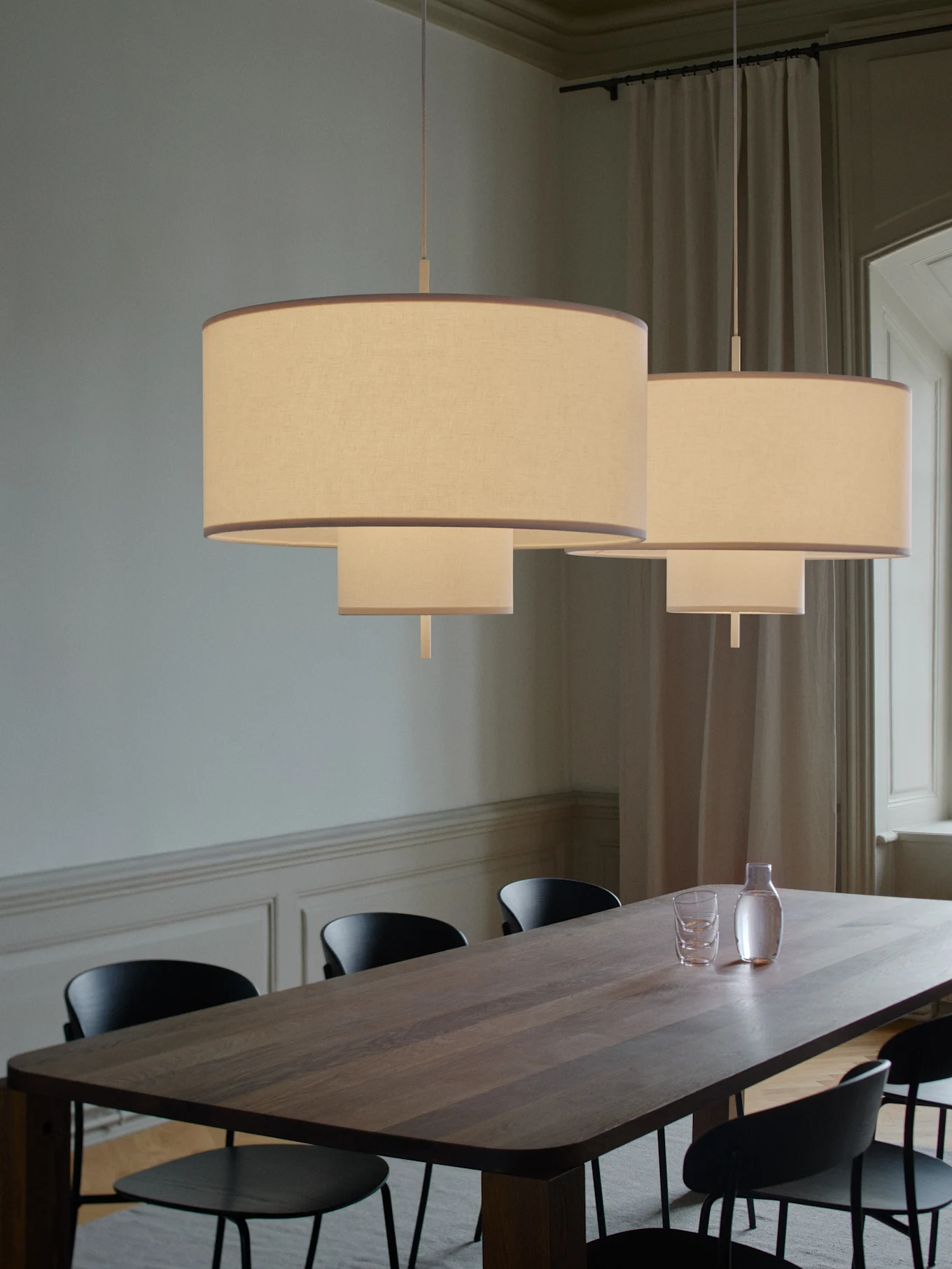 Twee gelaagde stoffen hanglampen verlichten een lange houten eettafel met zwarte stoelen, een karaf en glazen.