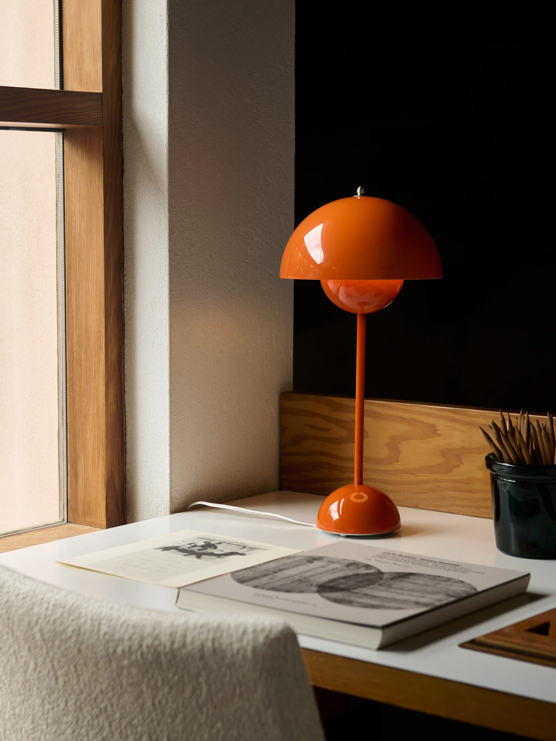 Een oranje koepelvormige tafellamp staat op een wit bureau naast boeken en een pennenbakje.