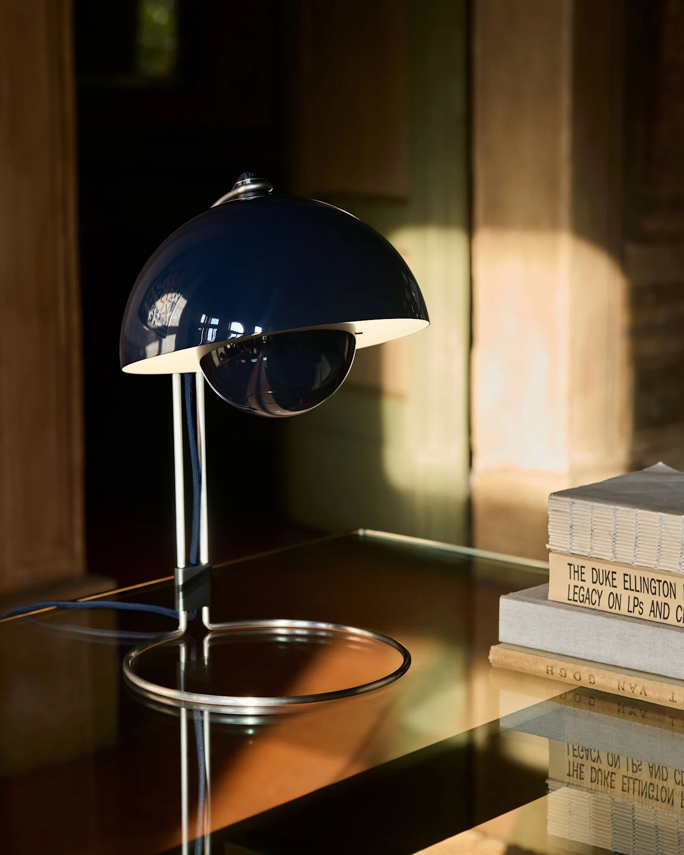Een donkerblauwe, klokvormige tafellamp met chromen voet staat op een reflecterende tafel naast een stapel boeken.