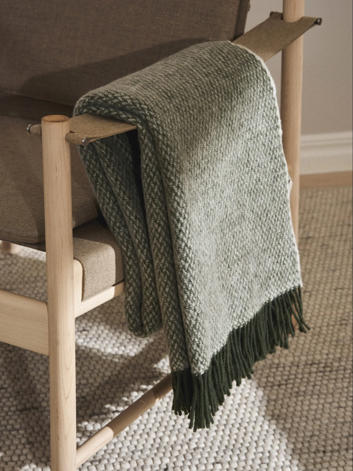 Een lichtbruine houten fauteuil met een groen en wit gedessineerde plaid over de armleuning, voorzien van donkergroene franjes.