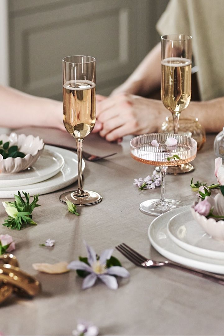 Je kunt de tafel ook op een speelse manier versieren voor je romantische diner thuis. Hier zie je wit servies, eenvoudige glazen en bloemen op een beige tafelkleed.