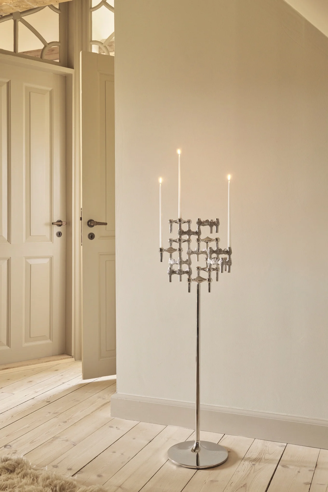 Hoge, moderne zilveren kandelaar met brandende witte kaarsen op een houten vloer in een kamer met lichte muren en open deuren.