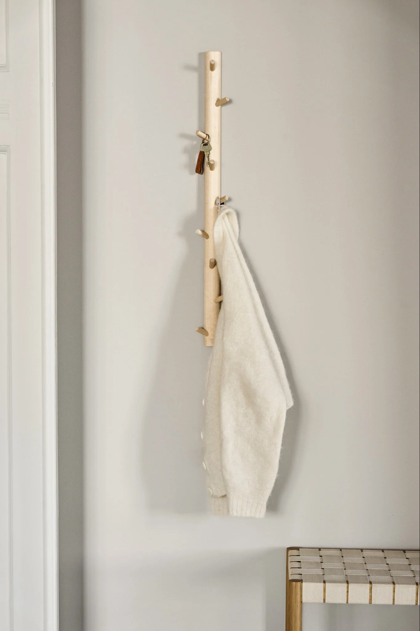 Een licht houten kapstok met haken houdt sleutels en een crèmekleurige trui, met daaronder een gevlochten bank.