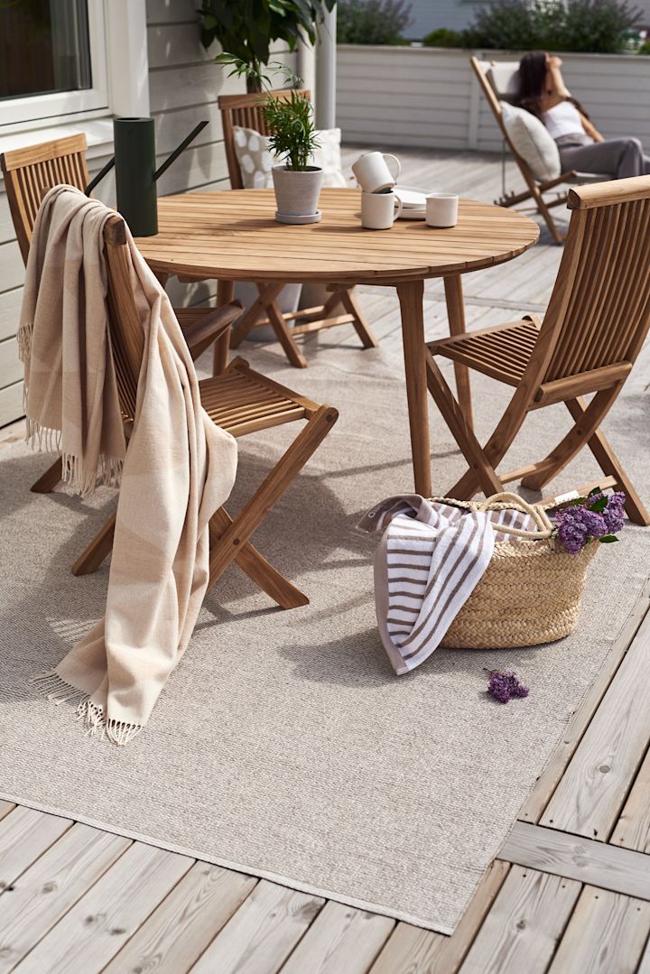 Een terras met houten meubels, bijvoorbeeld de Aneboda balkonset uit 1898 en het Rime katoenen vloerkleed van Scandi Living op het Mellow plastic tapijt.