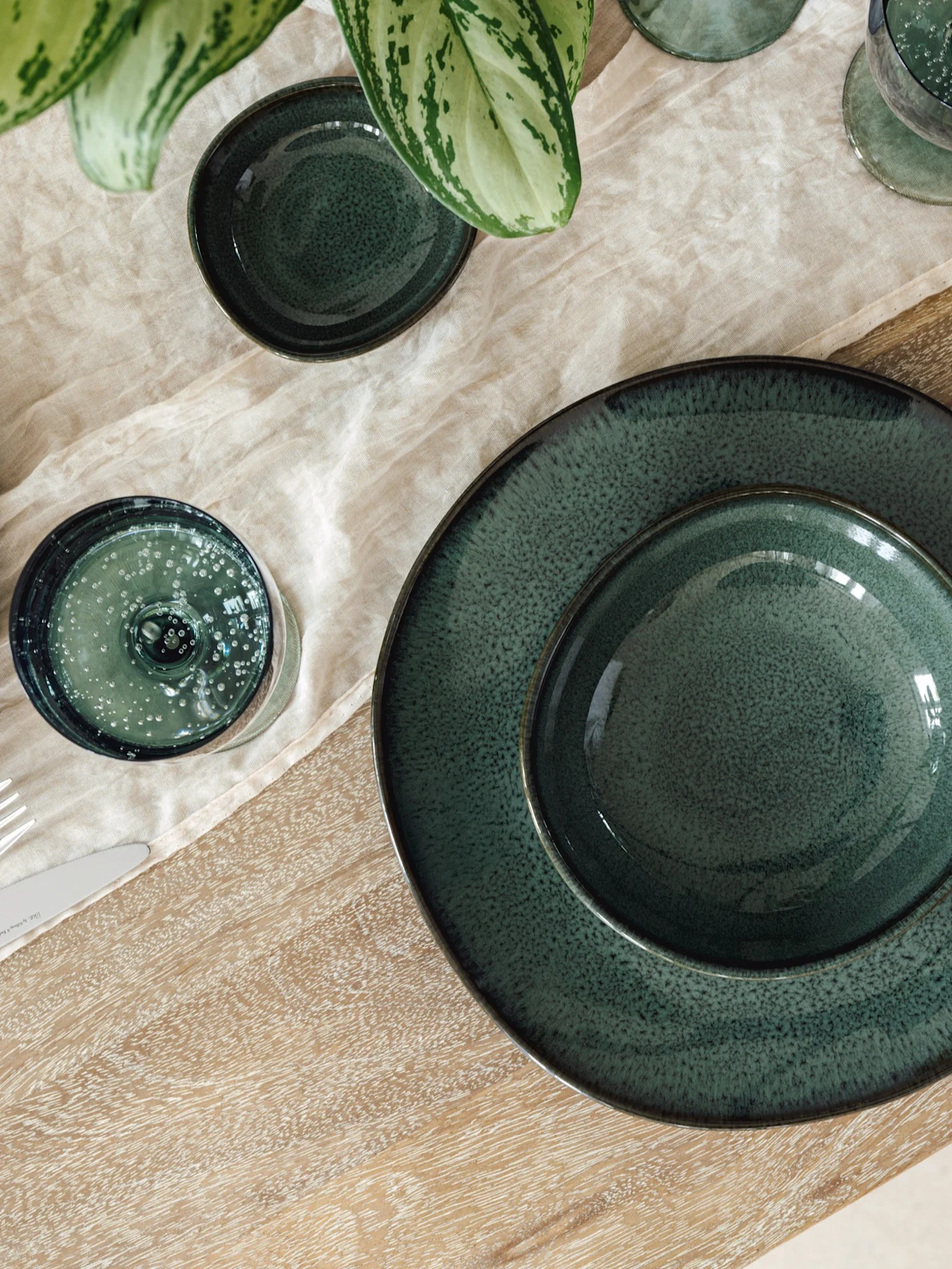 Gedekte tafel met donkergroene keramische borden, kommen en glazen op een lichte linnen loper en houten tafel, met groene plant.
