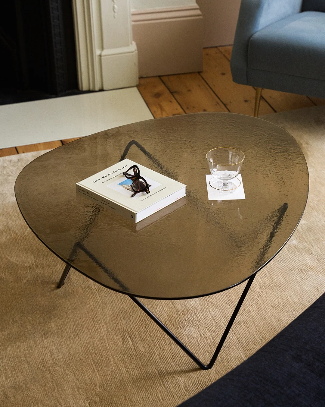 Organisch gevormde donkere glazen salontafel met een boek, een bril met schildpadmontuur en een glas met gouden rand op een onderzetter.