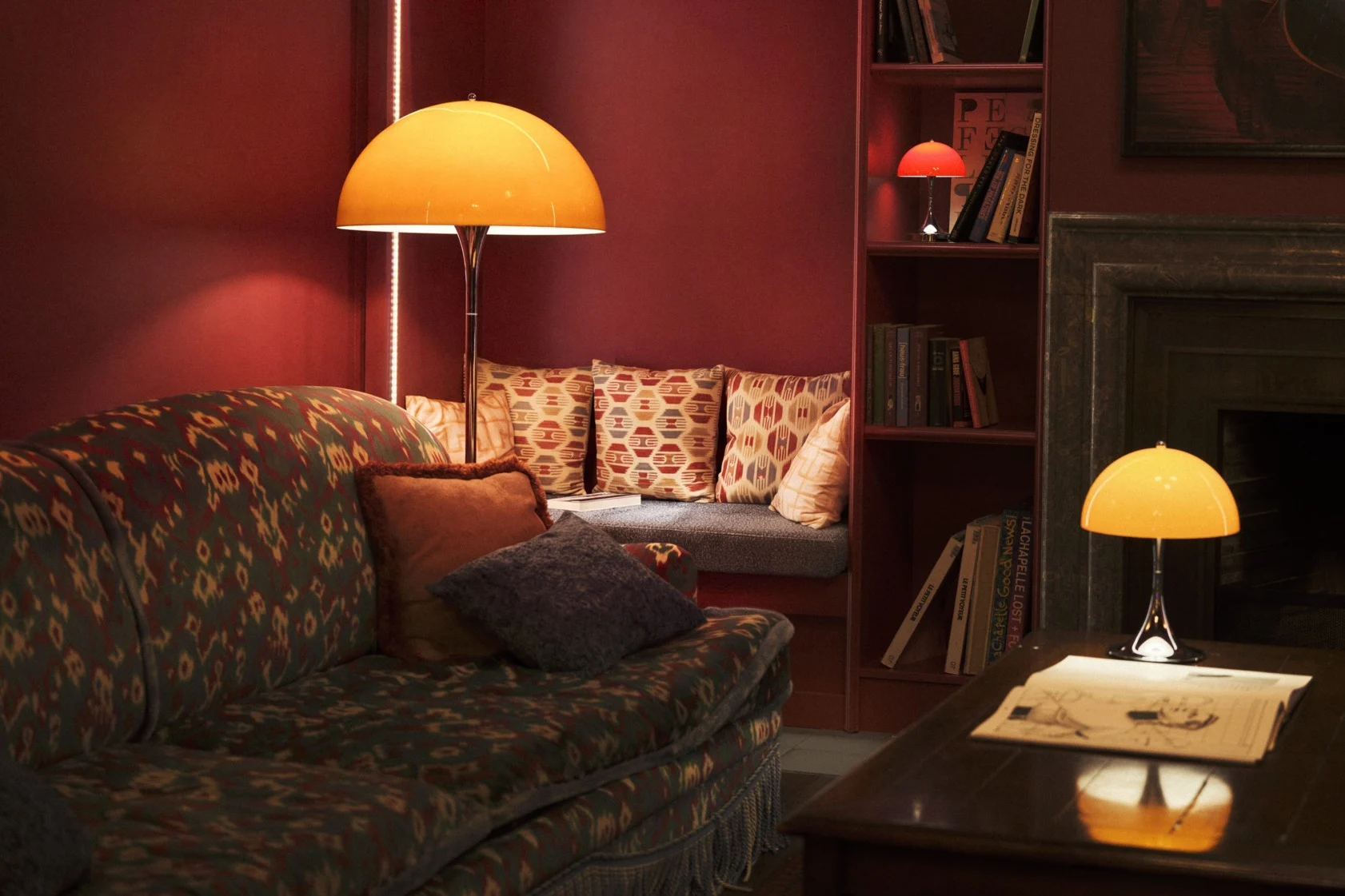 Een retro vloerlamp met oranje koepelkap en chromen standaard verlicht een kamer met een patroonkleed en houten kledingkast.