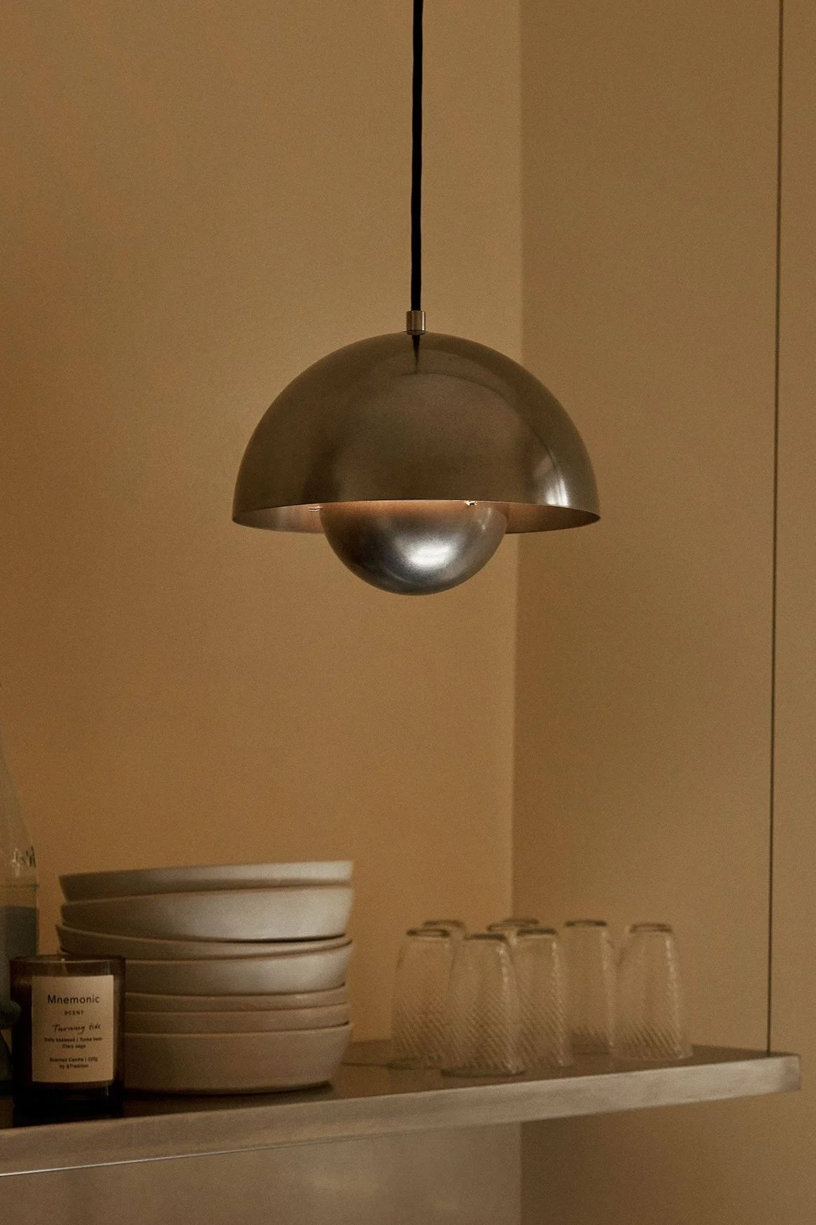 Een koepelvormige metalen hanglamp hangt boven een plank met kommen, glazen, flessen en een kaars tegen een warmbruine muur.