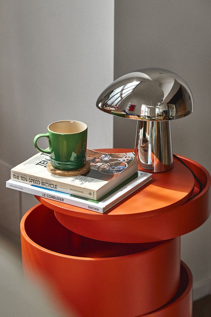 Geef je huis een update met deze moderne verlichtingsideeën zoals de Night Owl lamp van Fritz Hansen in chroom, hier te zien op een rode hoge tafel met wat boeken en een groen kopje. 