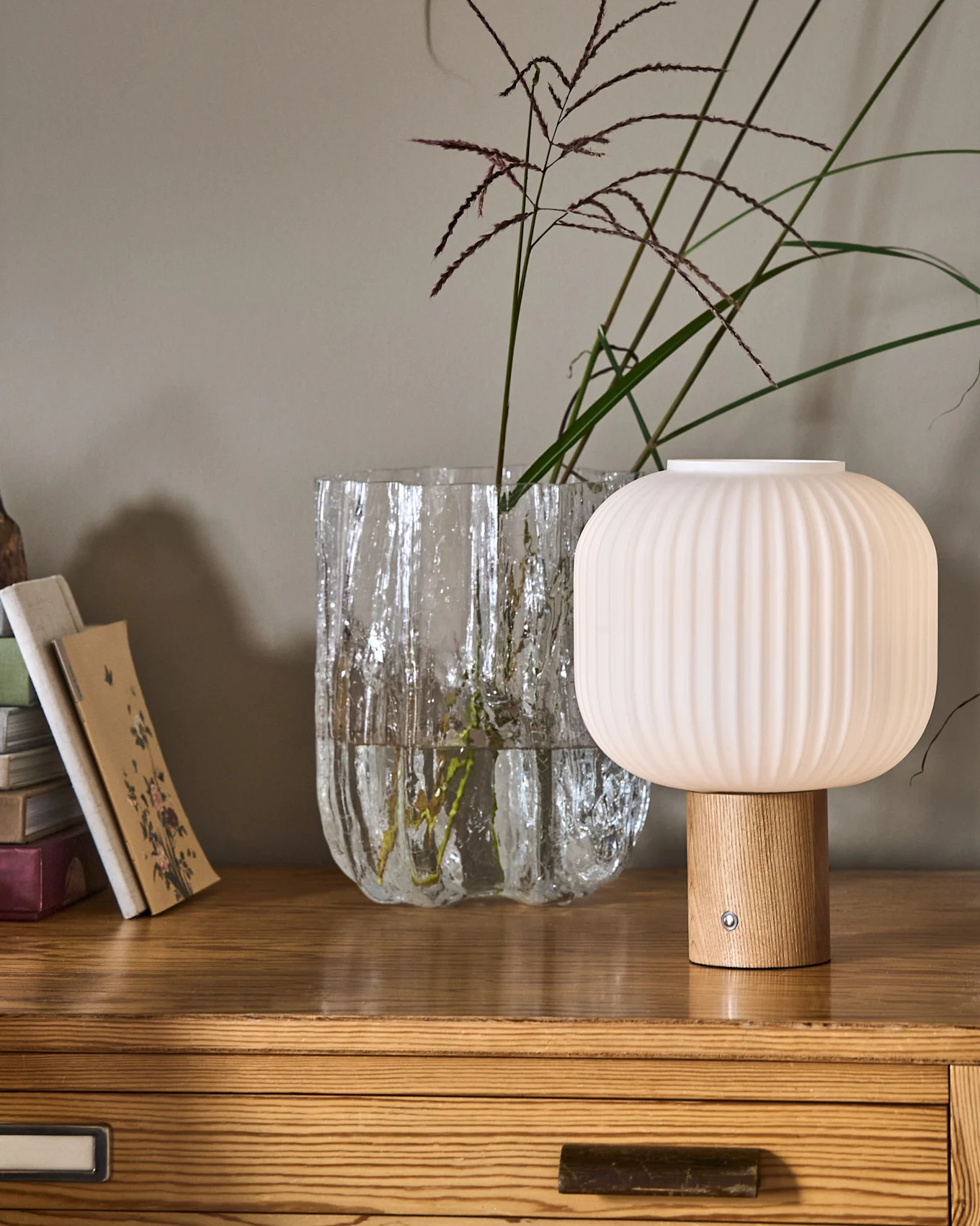 Een geribbelde, paddenstoelvormige lamp met houten voet naast een getextureerde glazen vaas met planten en een stapel boeken op een houten ladekast.