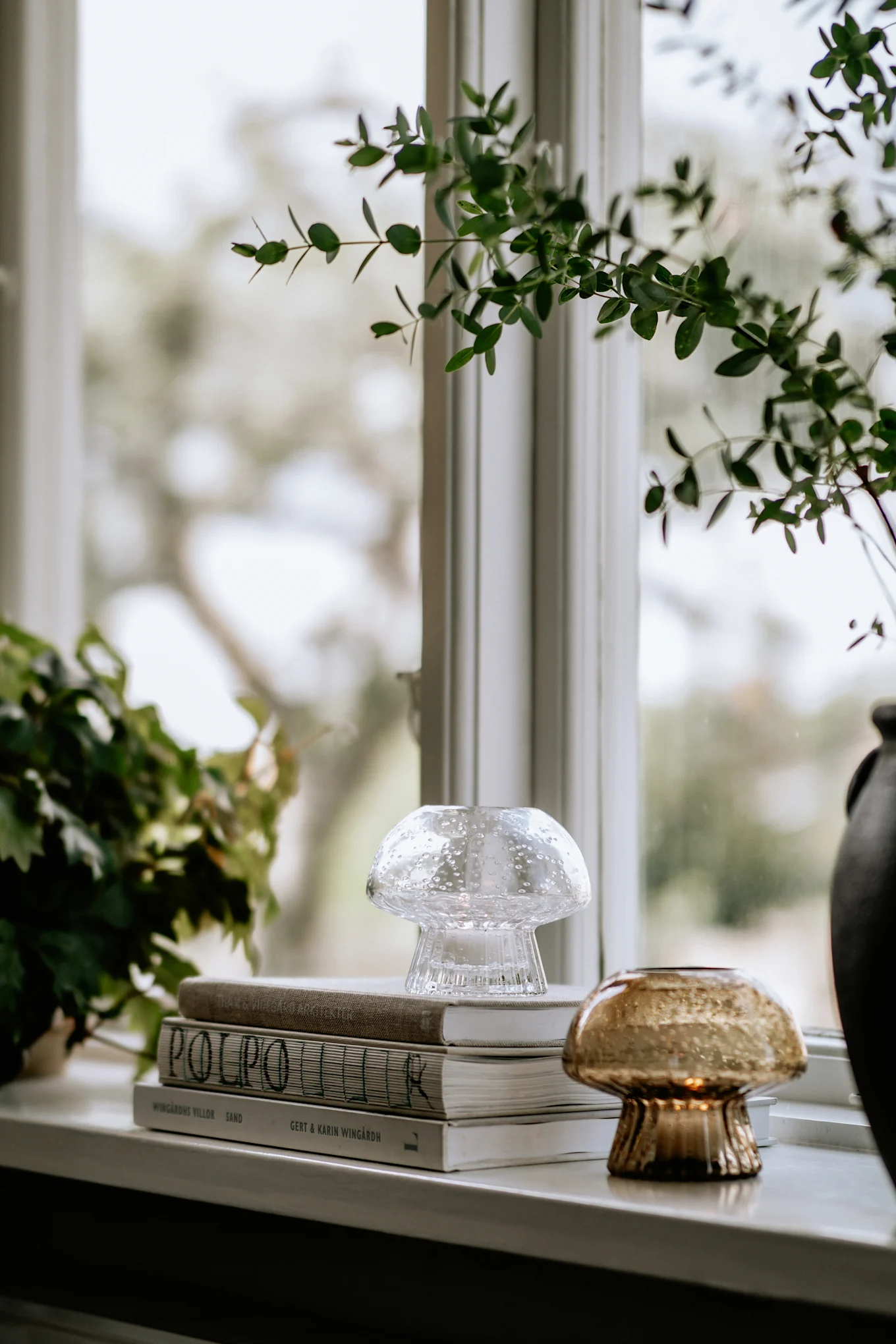 Transparante bubbelglas en amberkleurige theelichthouders, boeken en groene planten op een vensterbank.
