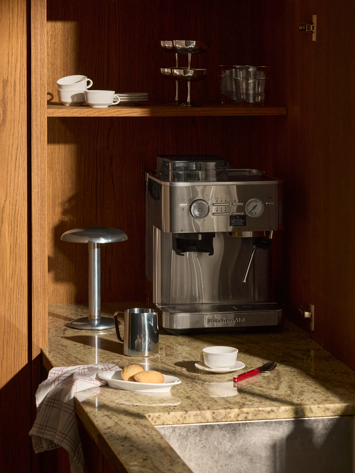 RVS espressomachine op granieten aanrecht in houten kast. Erboven kopjes, glazen. Op aanrecht koekjes, kopje, melkopschuimer.