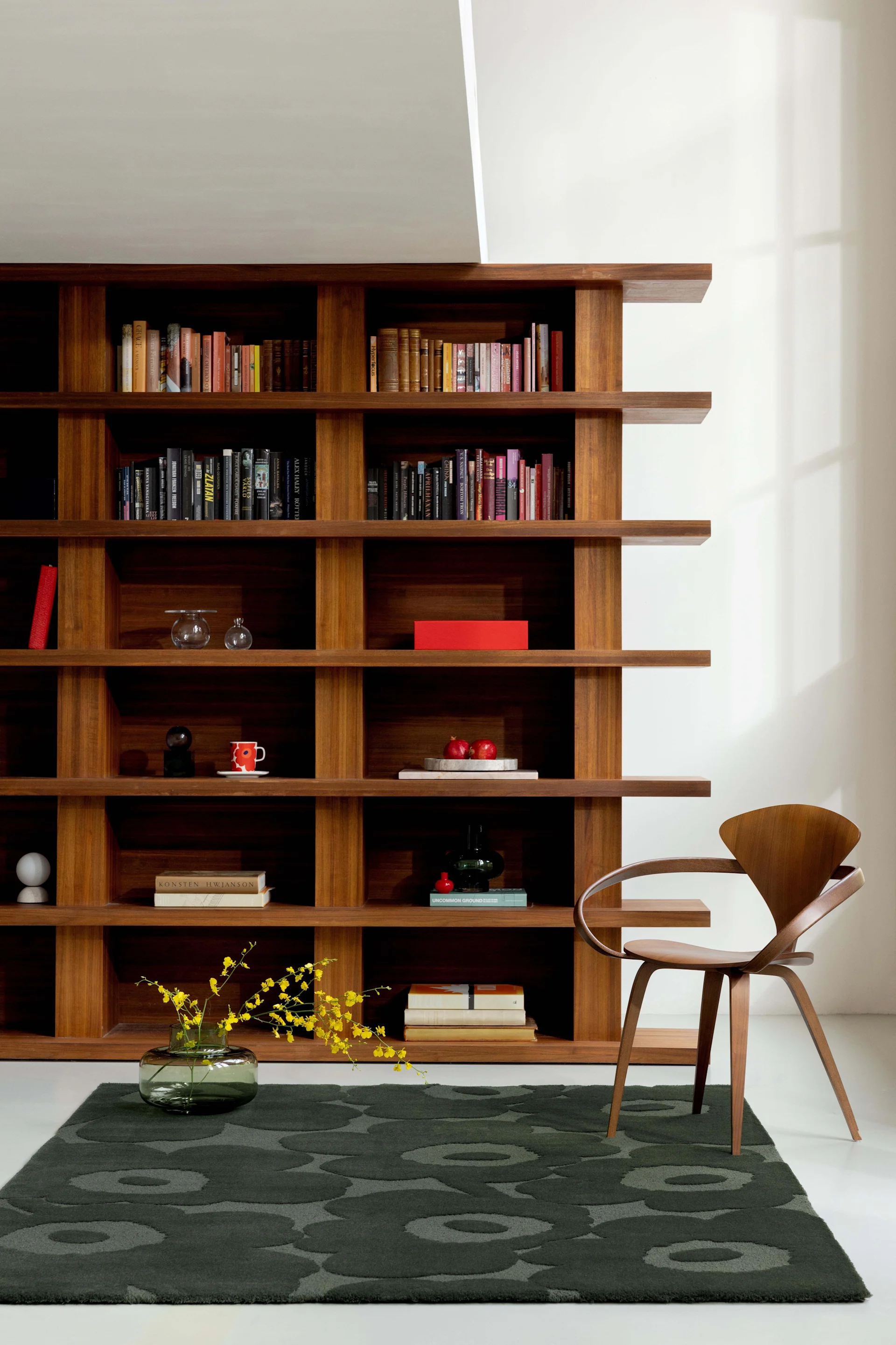 Een ruime woonkamer met een hoge houten boekenkast vol boeken en decoratie, een donkergroen bloemenkleed en een gebogen houten stoel.