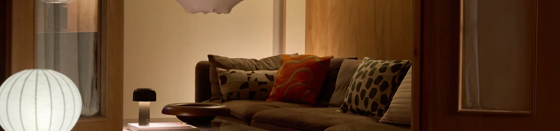 Een gezellige woonkamer met een bruine bank, diverse kussens met patronen en twee lampen: een grote witte geribbelde vloerlamp en een kleine paddenstoelvormige tafellamp.