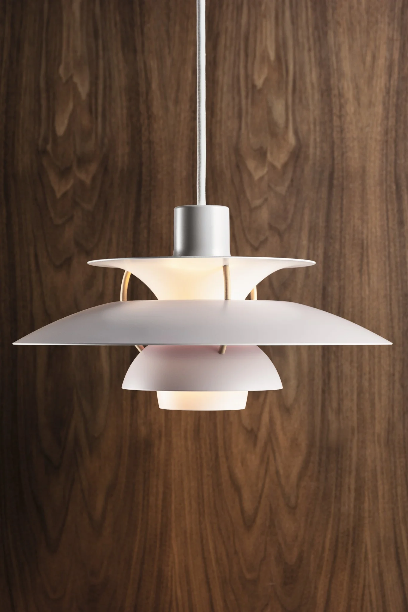 Een witte, meerlaagse hanglamp met een warme gloed, hangend tegen een donkere houten paneelwand.