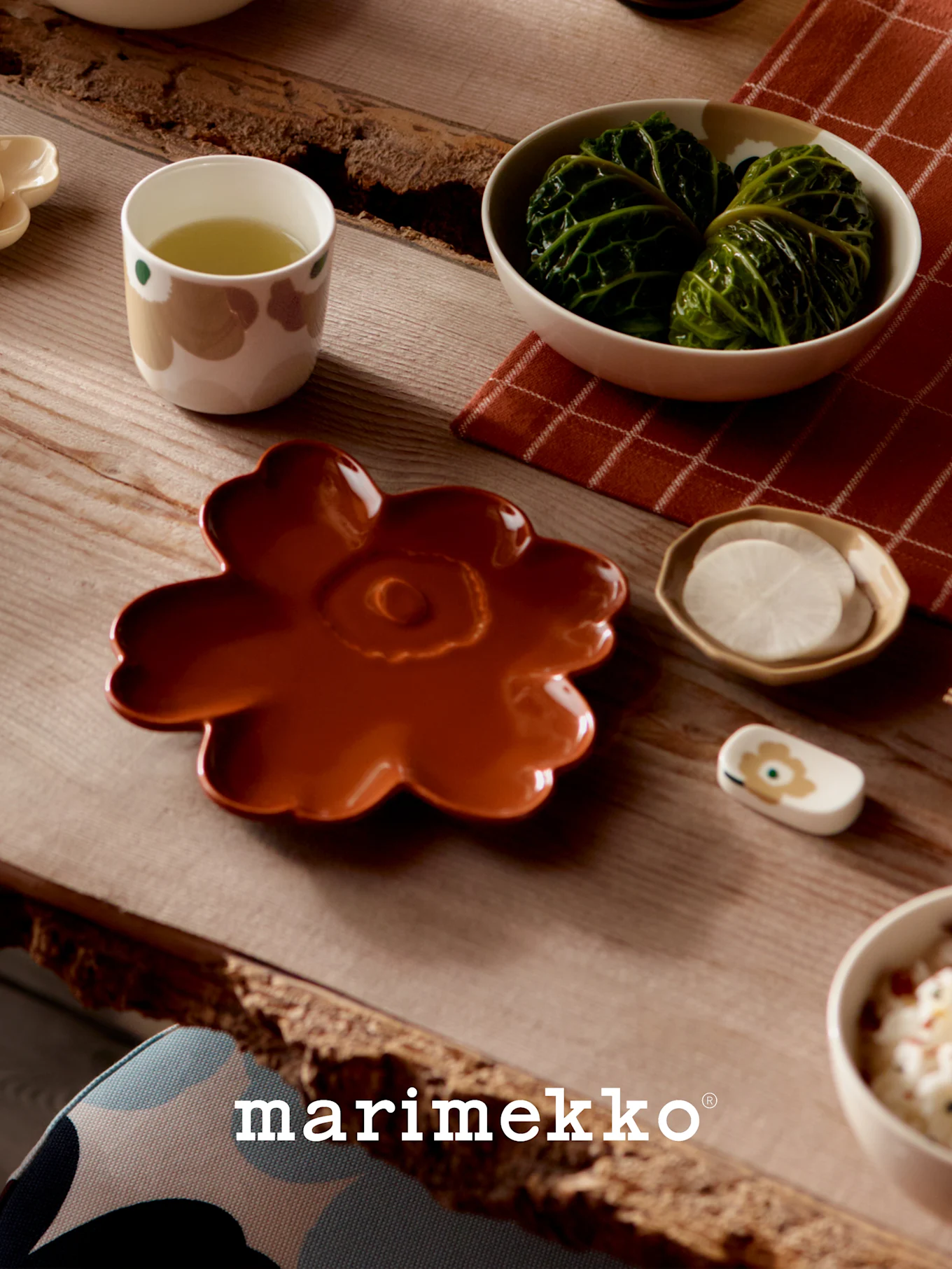 Tafel gedekt met Marimekko servies: een roestkleurig bloemenbord, een kop groene thee, koolrolletjes en gesneden radijsjes.