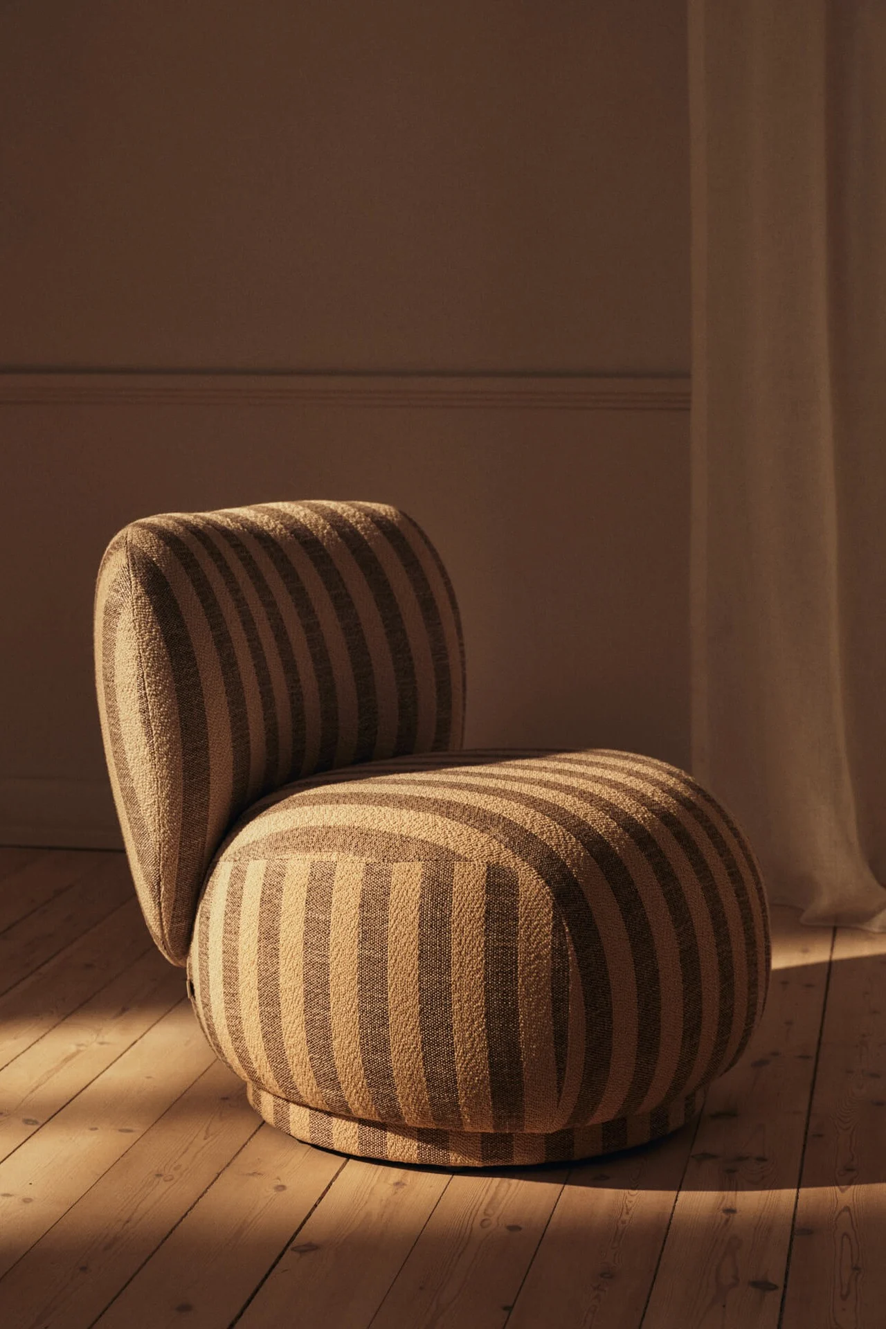 Moderne, afgeronde fauteuil met beige en bruine strepen op een houten vloer, badend in warm licht.
