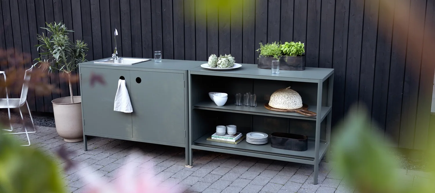 Een grijsgroene buitenkeukenunit, met een spoelbak en handdoek links, en planken met keukengerei en potkruiden rechts.