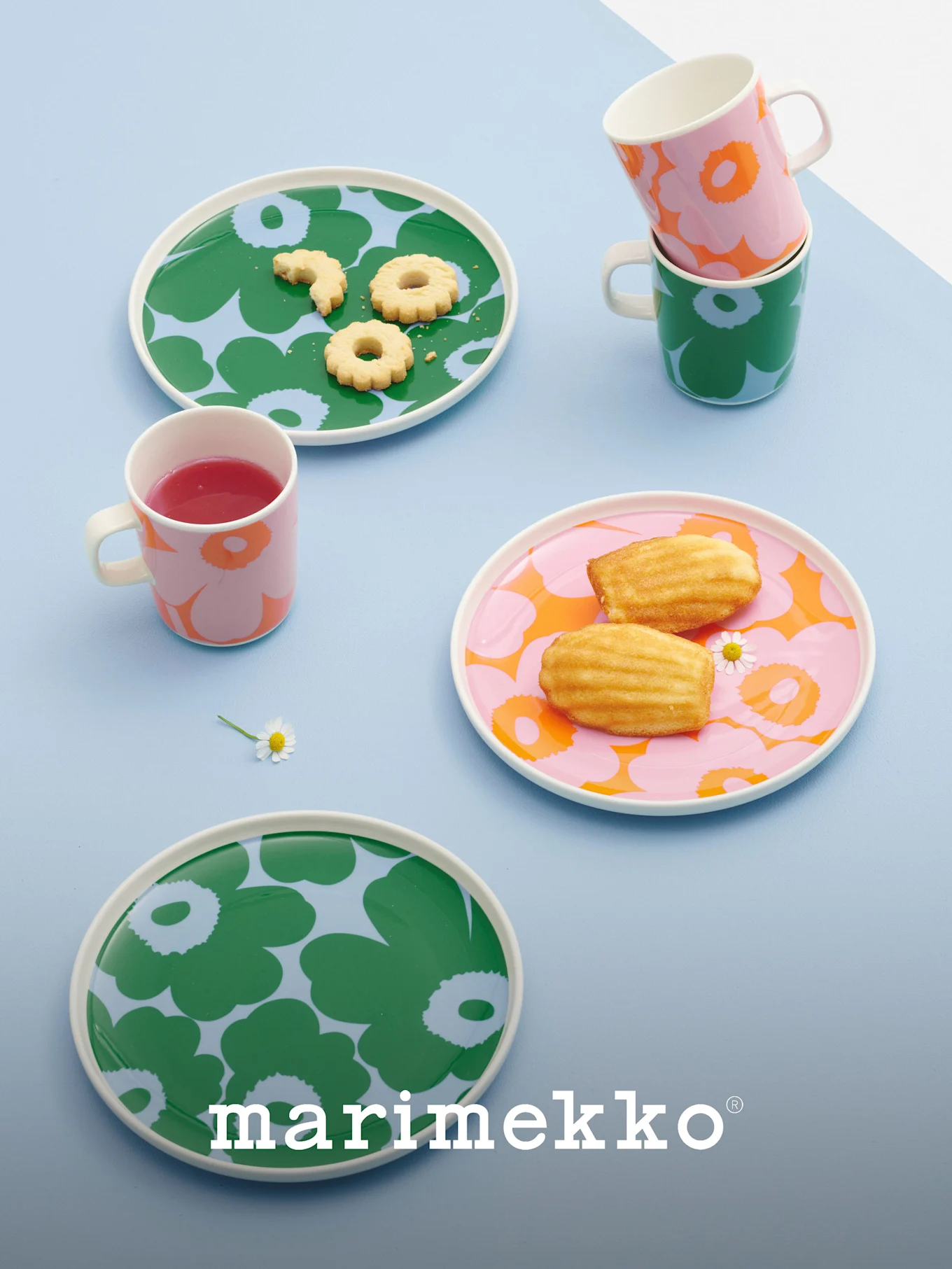 Marimekko borden en mokken met gedurfde bloempatronen in groen, roze en oranje, met koekjes, madeleines en een drankje.