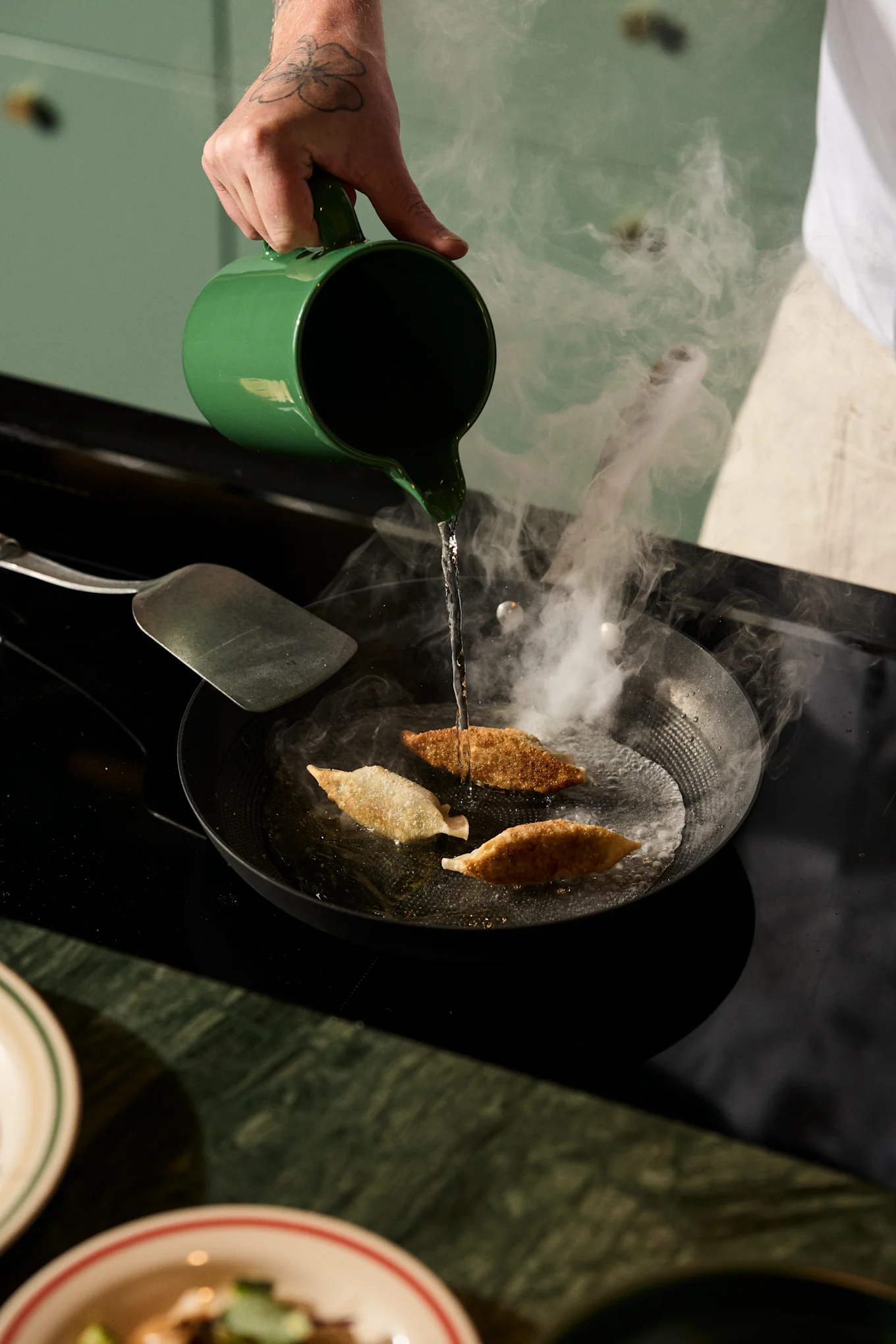 Een getatoeëerde hand giet water uit een groene kan in een hete pan met drie bakende dumplings, wat stoom creëert.