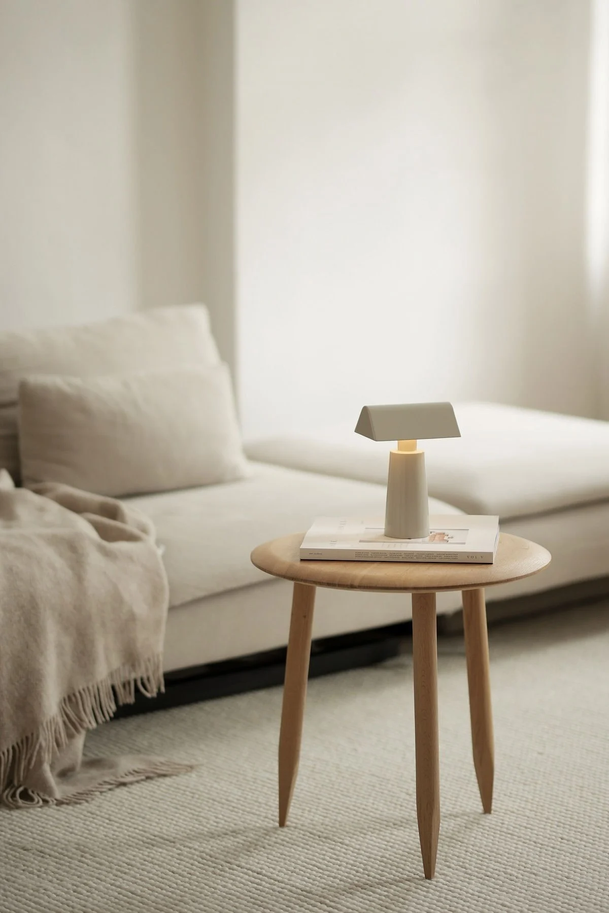 Een minimalistische lichtgrijze draagbare lamp met een rechthoekige kap op een ronde lichte houten bijzettafel naast een beige bank met een plaid.