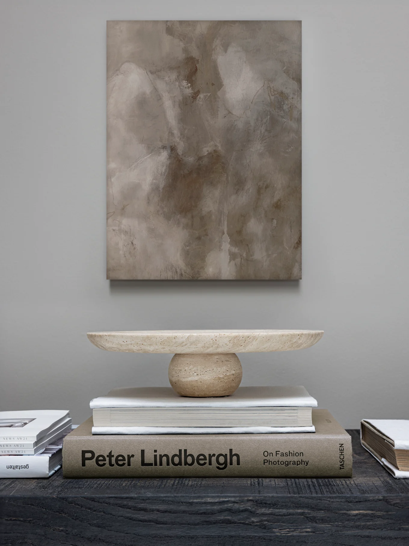 Een abstract schilderij hangt boven een lichte stenen decoratieve schaal op een stapel boeken, waaronder 'Peter Lindbergh', op een donkere tafel.