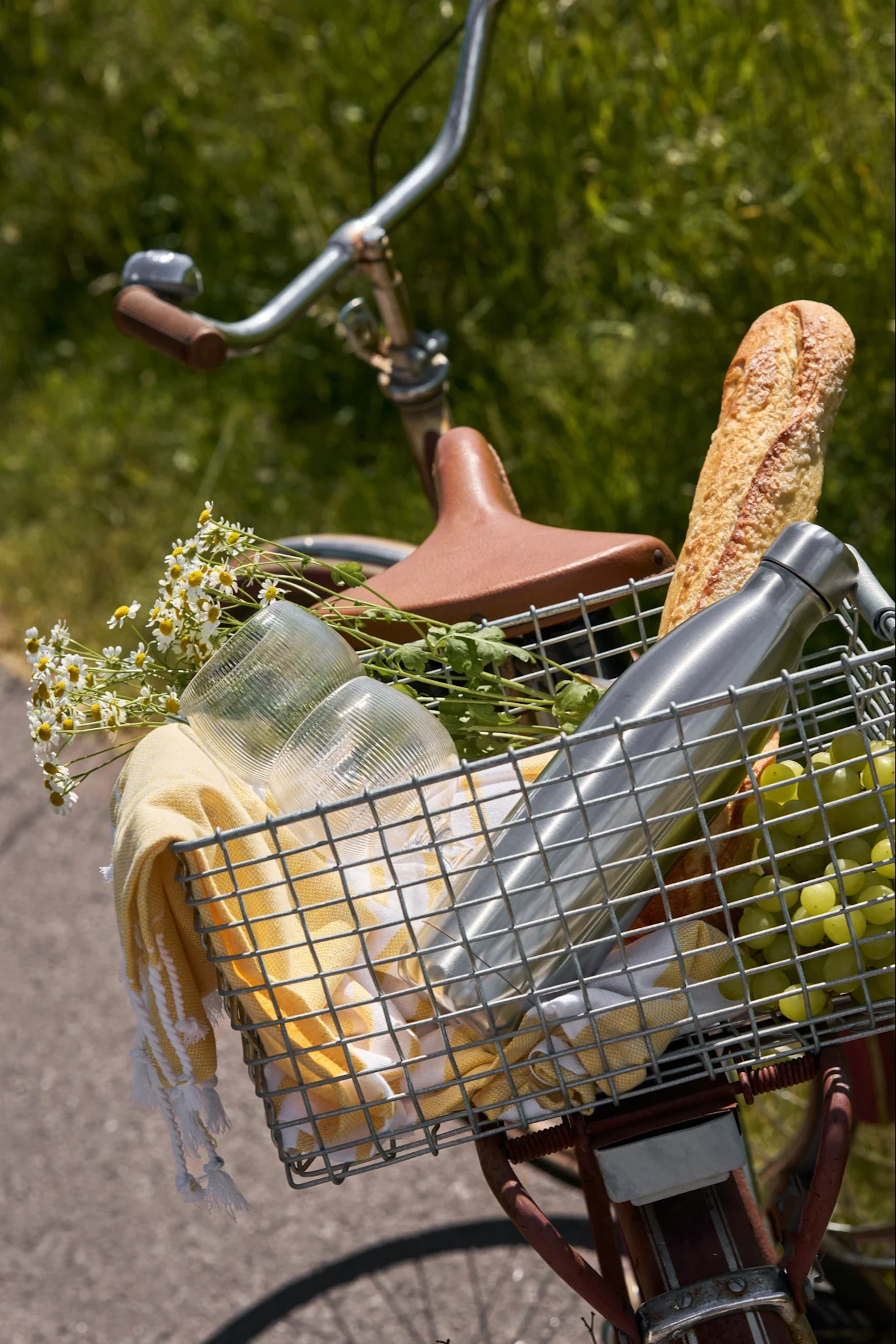 Fietsmand gevuld met picknickartikelen: een baguette, waterfles, druiven, glazen, gele handdoek en witte madeliefjes.