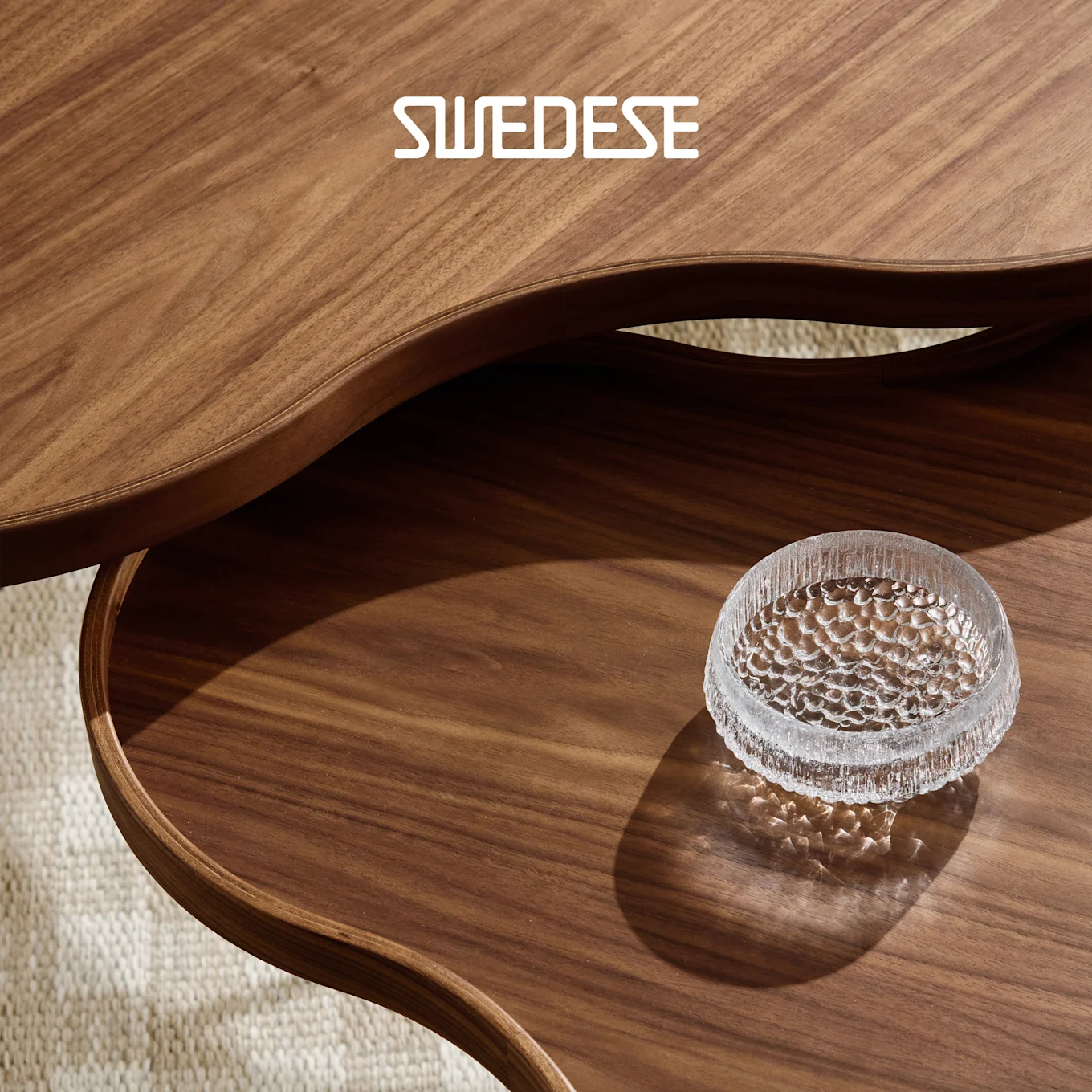 Twee gebogen walnoothouten tafels, de een de ander overlappend, met een getextureerde heldere glazen kom op de onderste tafel. SWEDESE-logo.