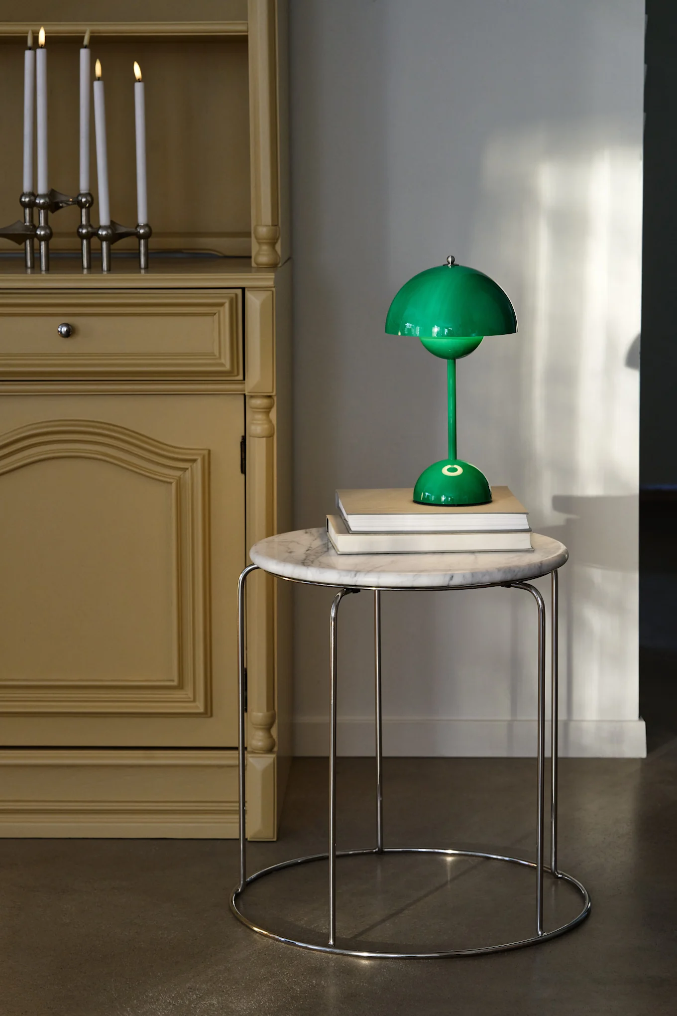 Een groene paddestoelvormige tafellamp staat op boeken op een ronde marmeren tafel, naast een gele kast met brandende kaarsen.