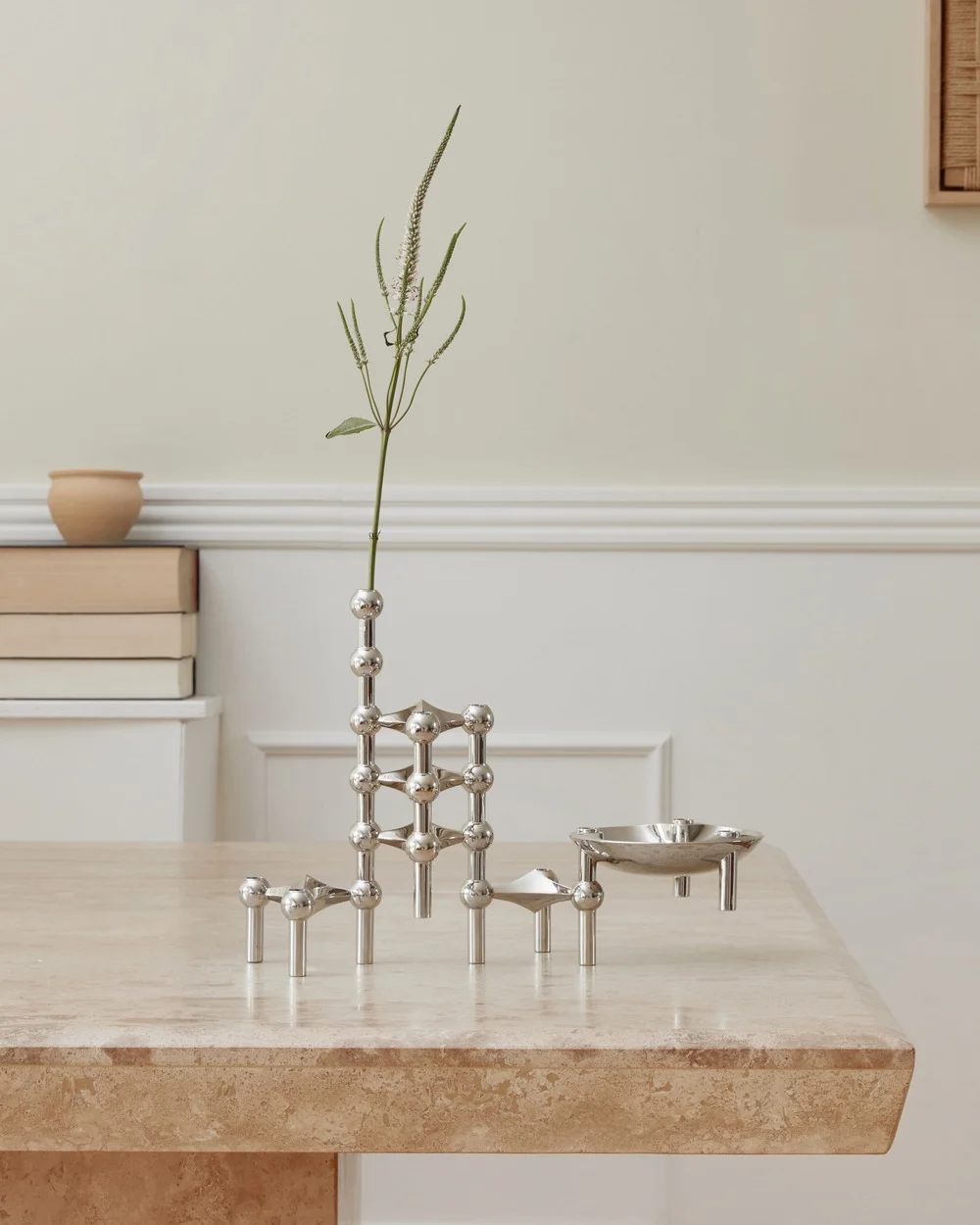 Een gepolijst zilveren modulair kandelaarsysteem met een plantenstengel en een klein schaaltje op een travertin tafel.