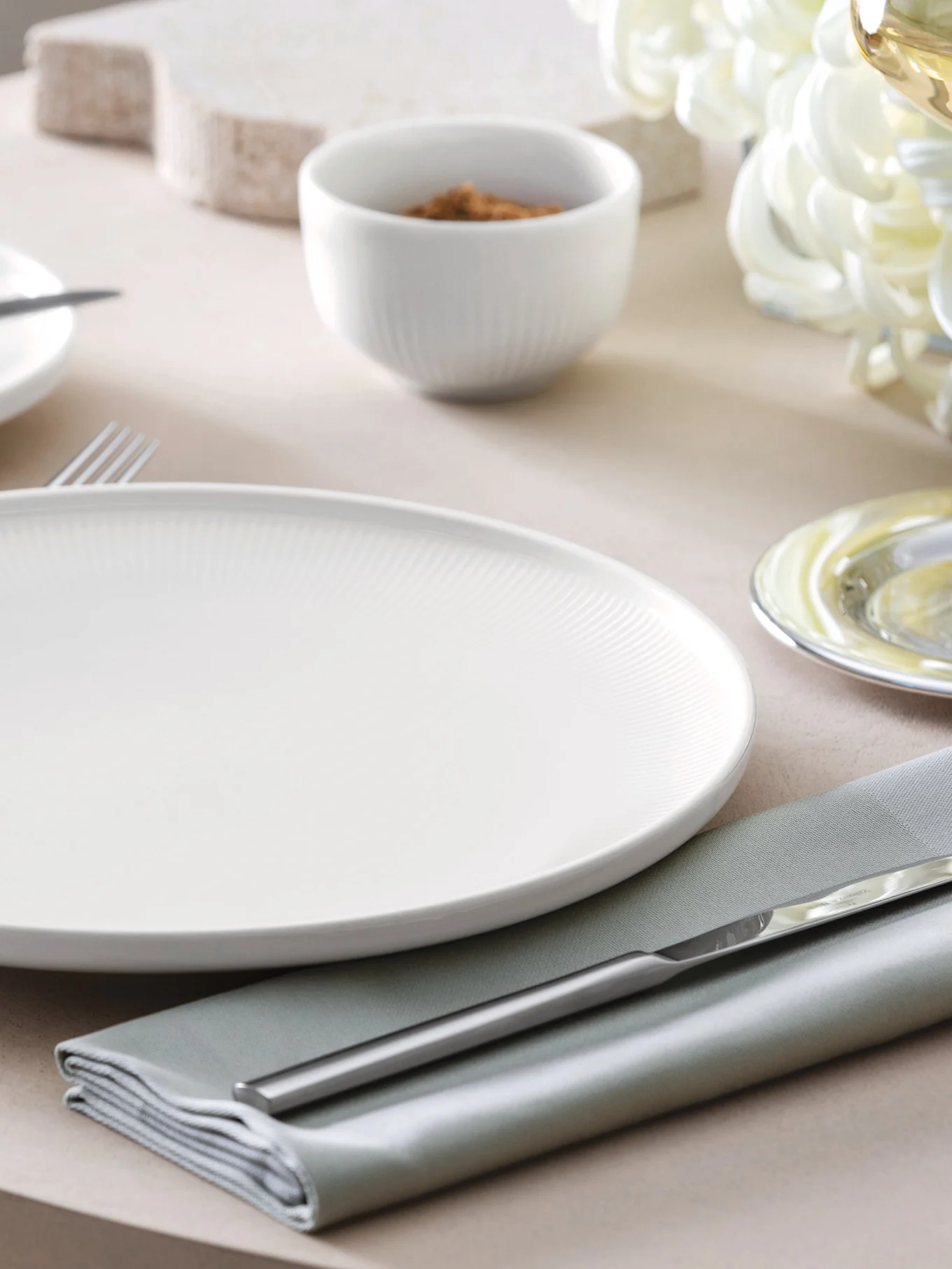 Een wit geribbeld dinerbord, saliegroen servet en zilveren bestek op een tafel met een klein kommetje op de achtergrond.