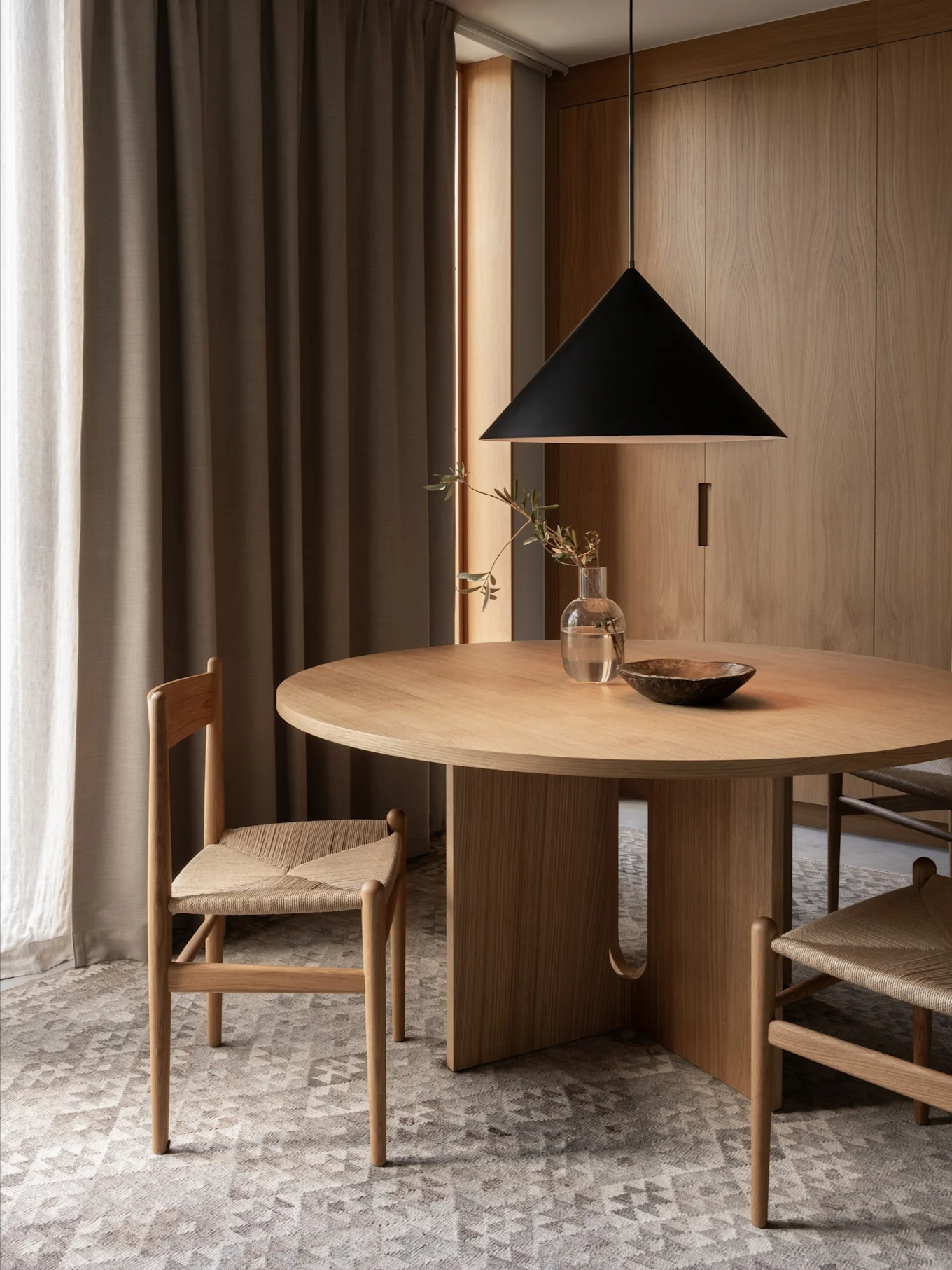 Een minimalistische eethoek met een ronde houten tafel, twee stoelen met gevlochten zittingen, een zwarte hanglamp en houten wanden.