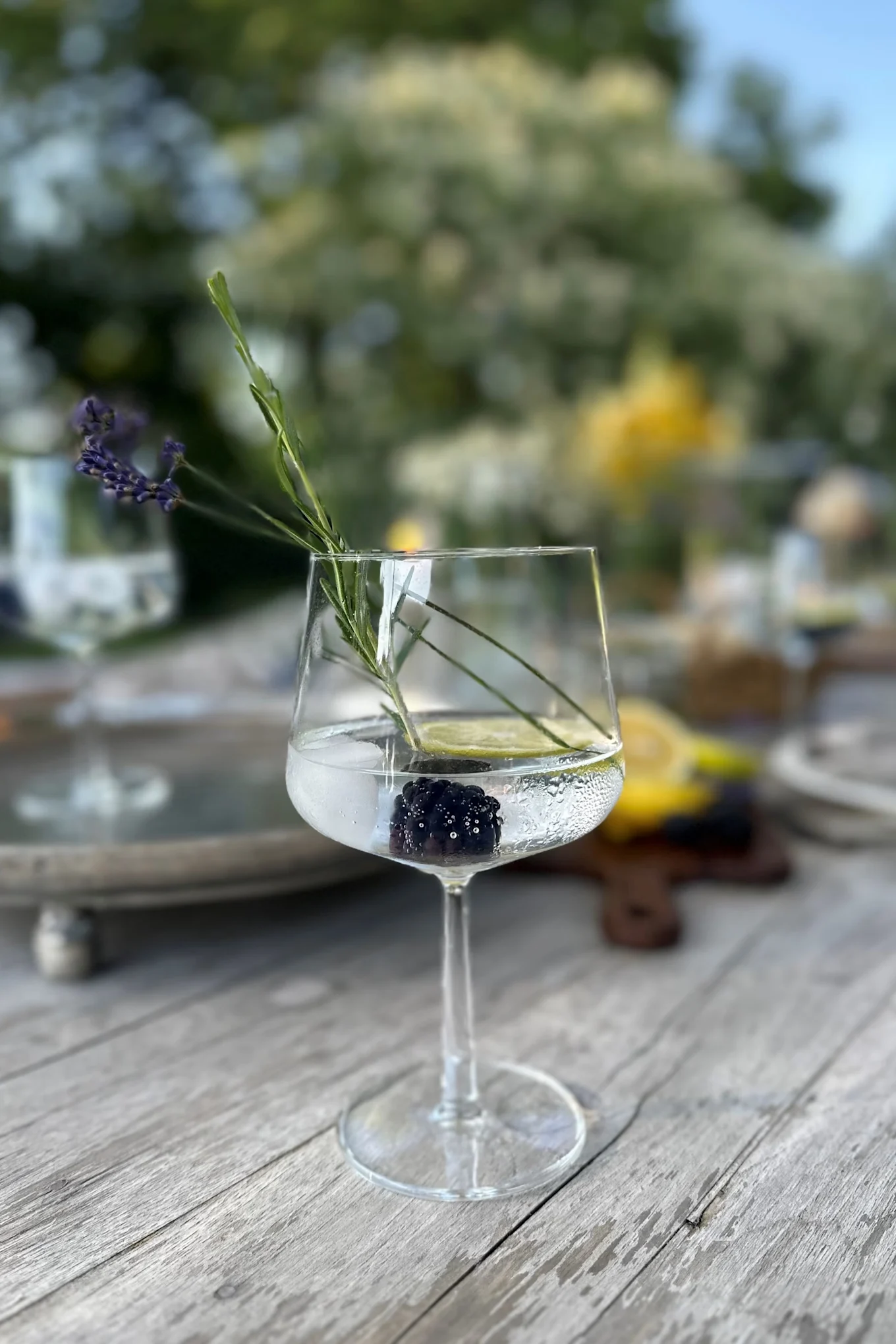 Een helder wijnglas op een houten tafel, met daarin een braam, een schijfje limoen, een takje lavendel en heldere vloeistof.