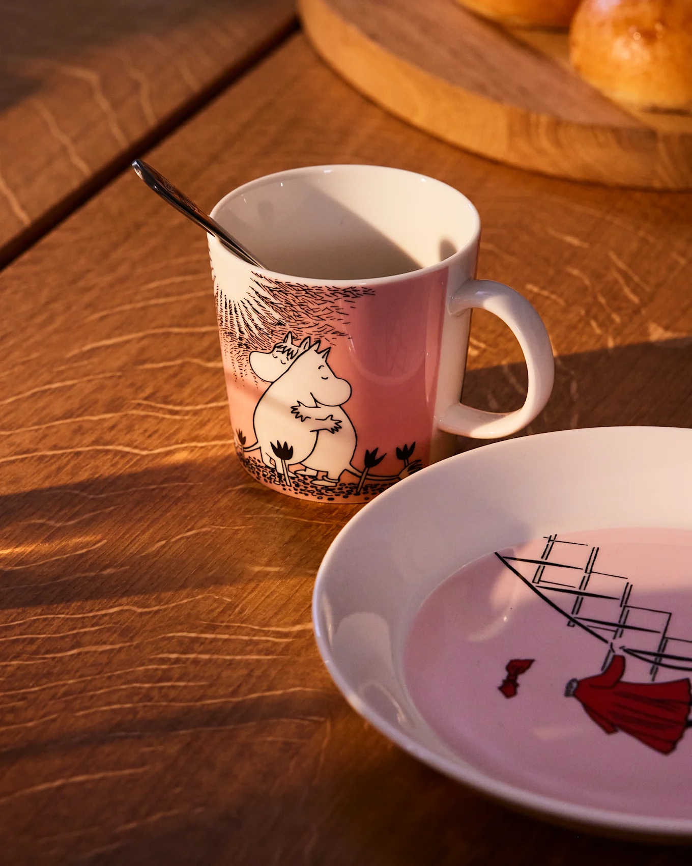 Roze Moomin-mok met Moomintrol en Snorkmeisje, naast een bijpassend bord met een zeilschip, op een houten tafel.