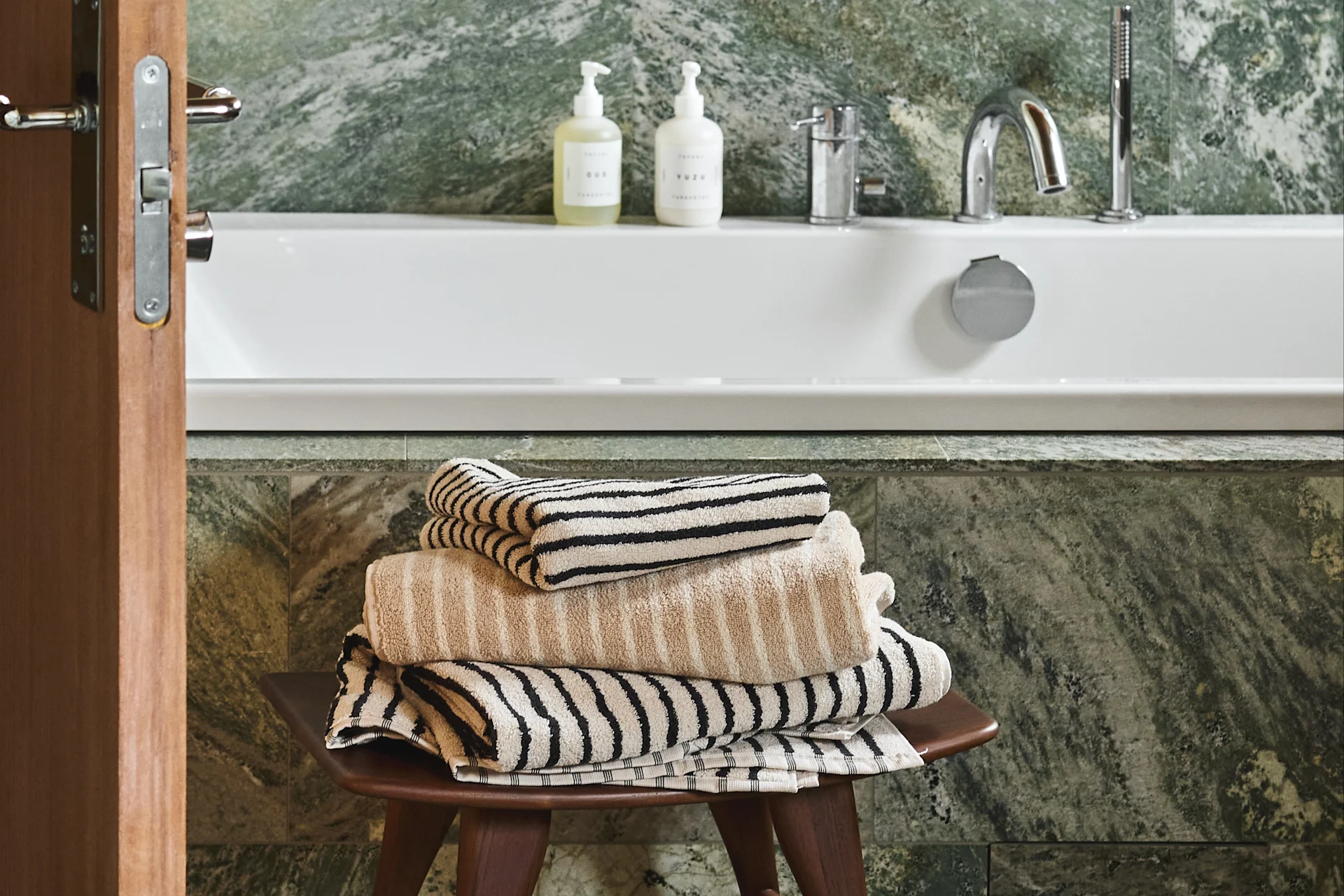 Een stapel beige en gestreepte handdoeken op een houten krukje in een badkamer met een wit bad en groene marmeren muren.