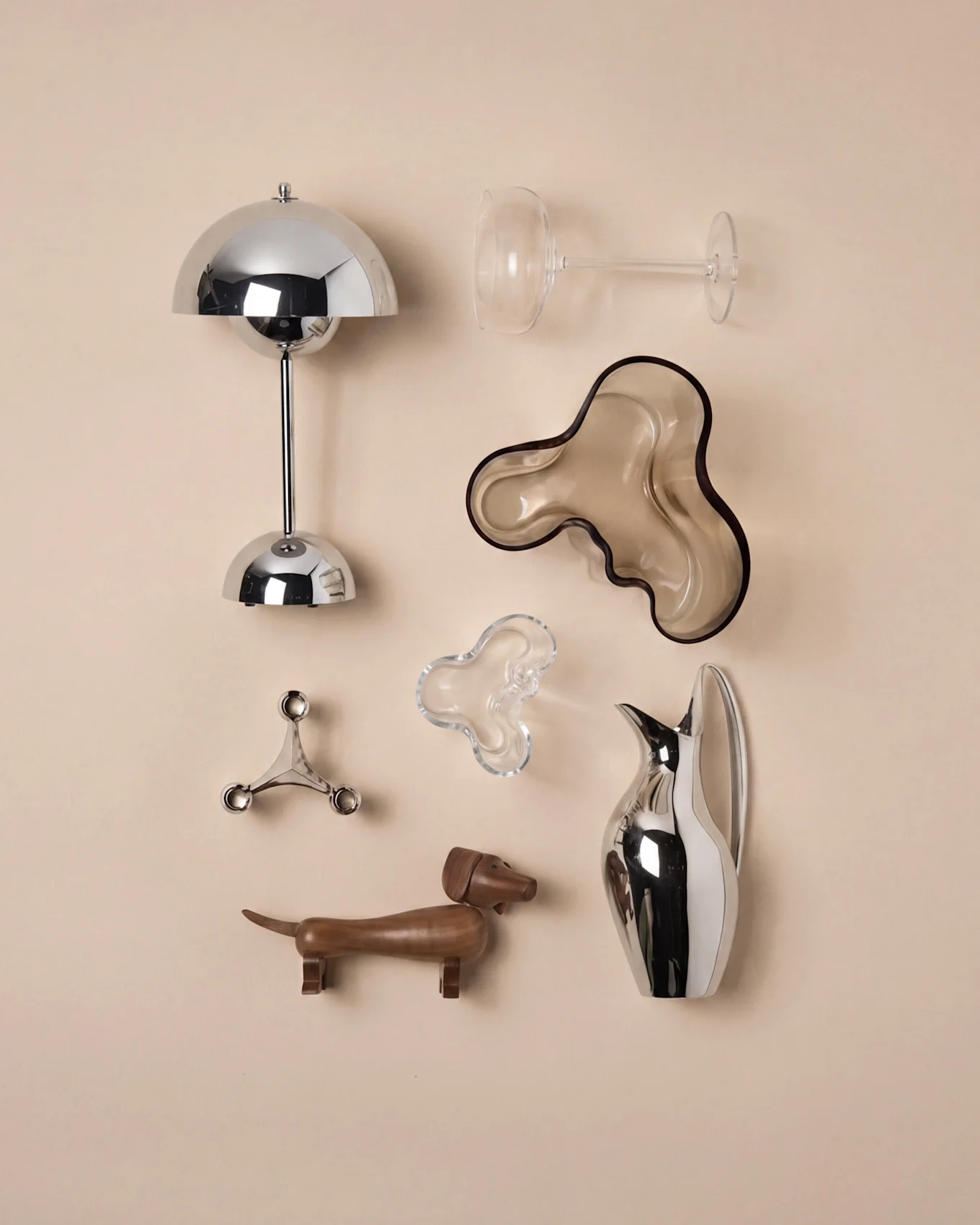 Moderne decoratie: chromen paddestoellamp, helder wijnglas, bruine golvende kom, zilveren kandelaar, houten teckel, zilveren kan.
