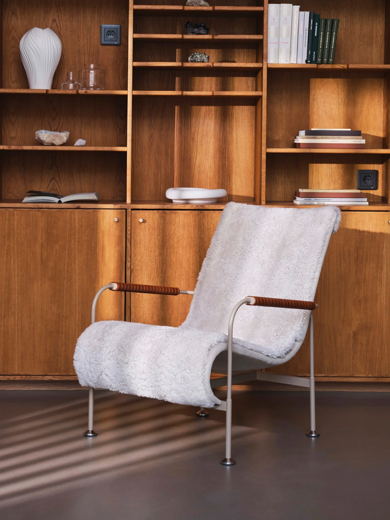 Witte kunstbont fauteuil voor een houten wandmeubel met planken, boeken, vazen en mineralen.